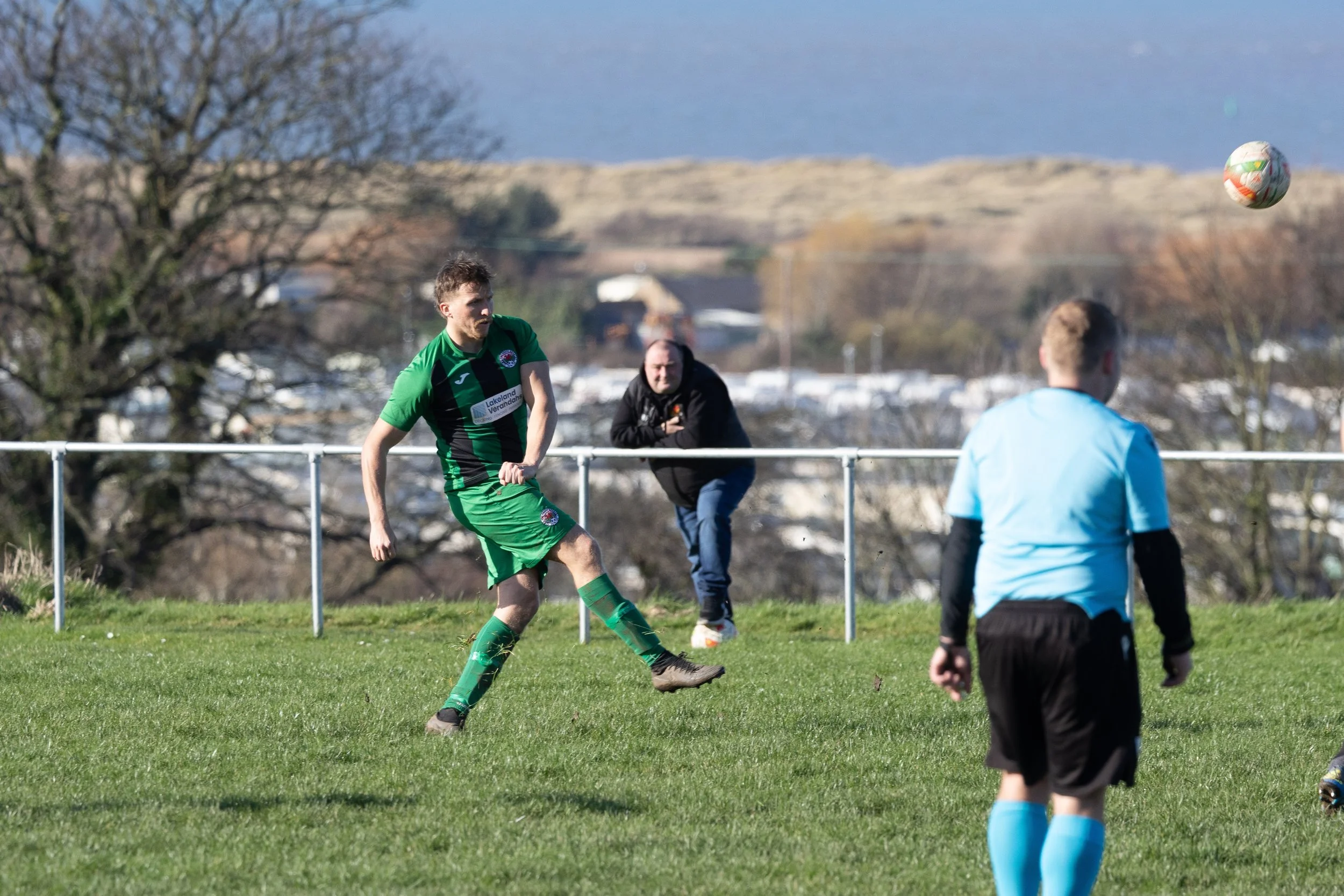 A soccer player in a green and black uniform is kicking a ball on a grassy field, with another player in a light blue uniform and a referee nearby, and a man watching in the background.