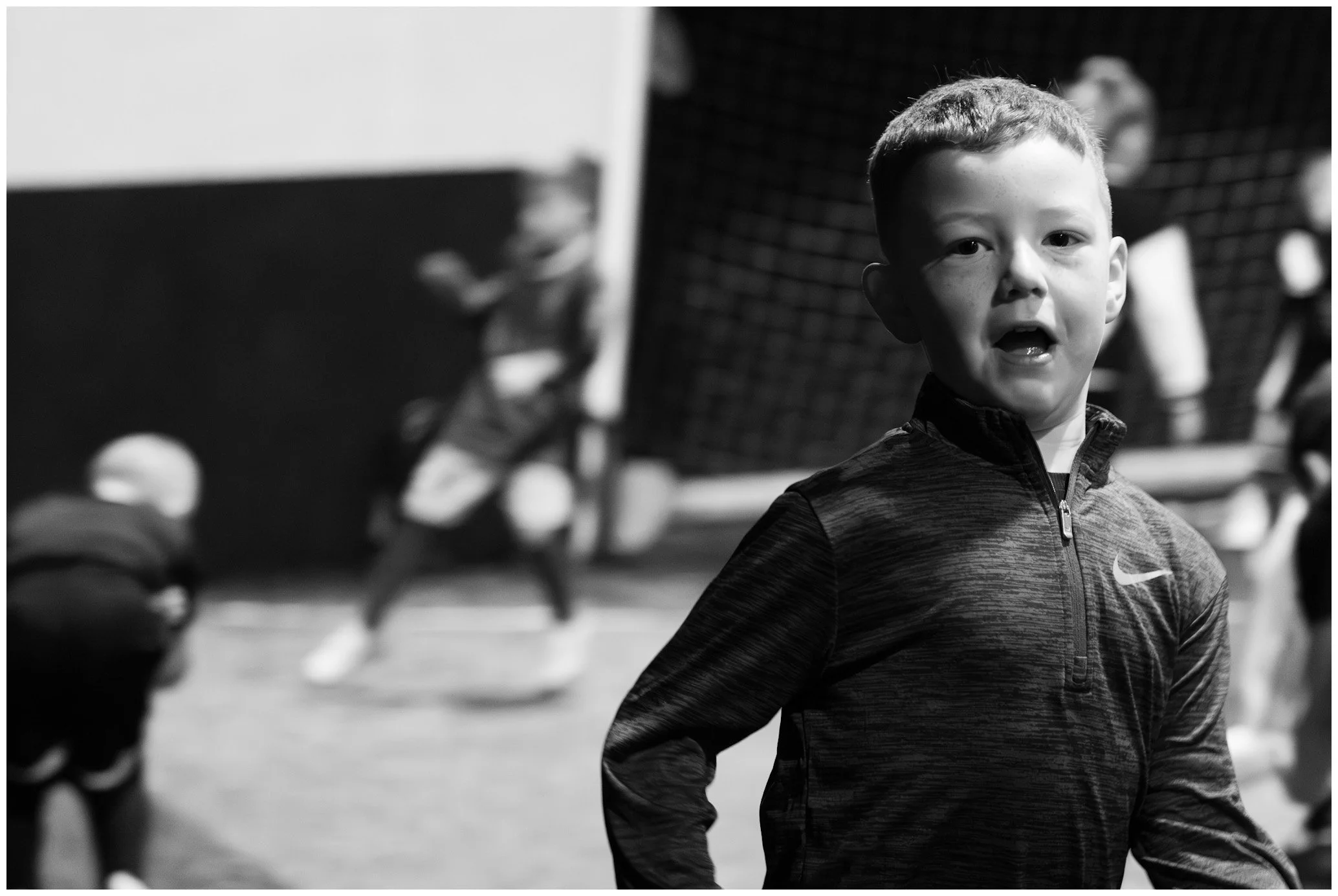 A young boy with short hair and a sporty jacket standing in an indoor sports facility, with other children blurred in the background playing basketball.