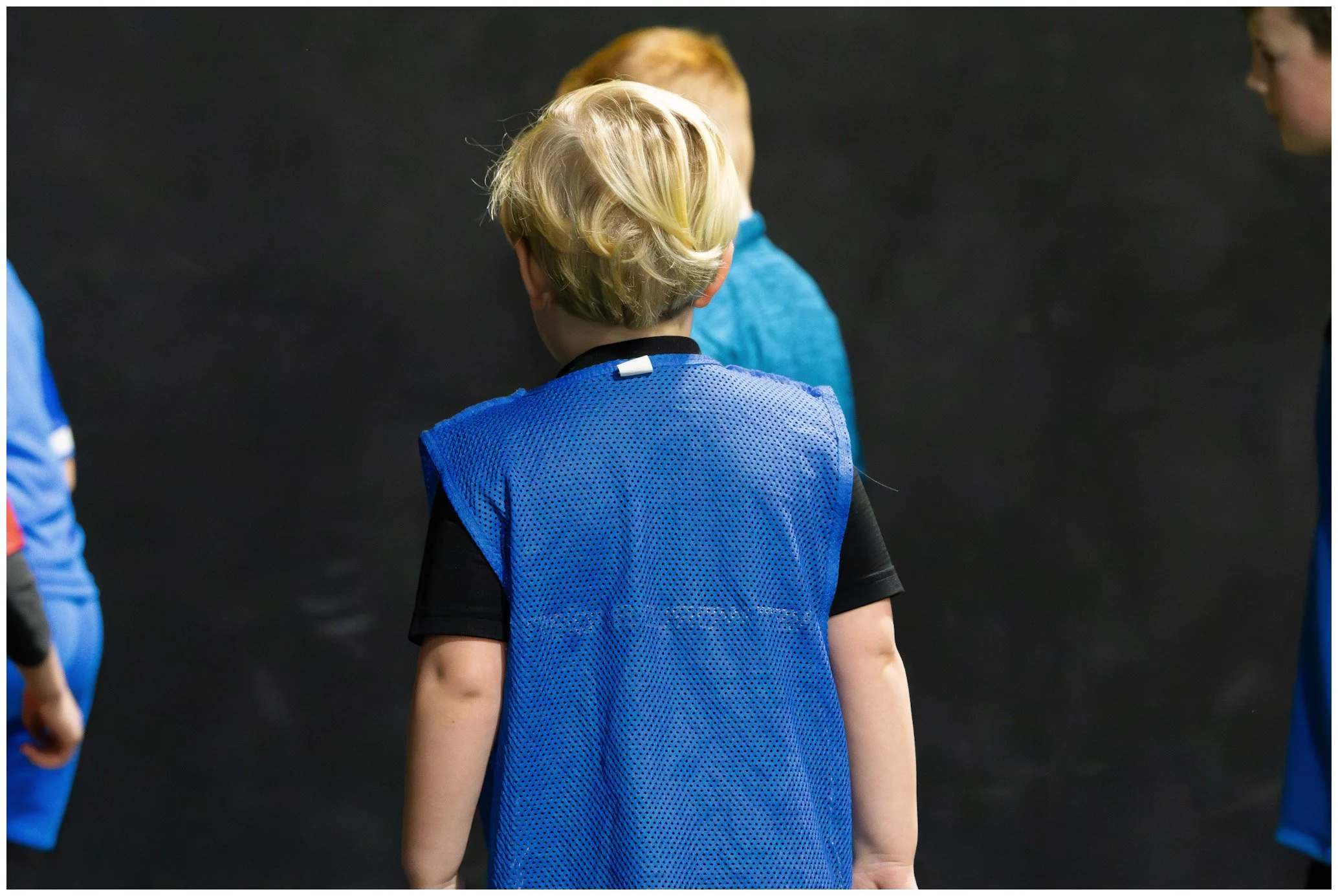 Child with blonde hair wearing a blue sports vest on a football field, facing away, standing among other children.
