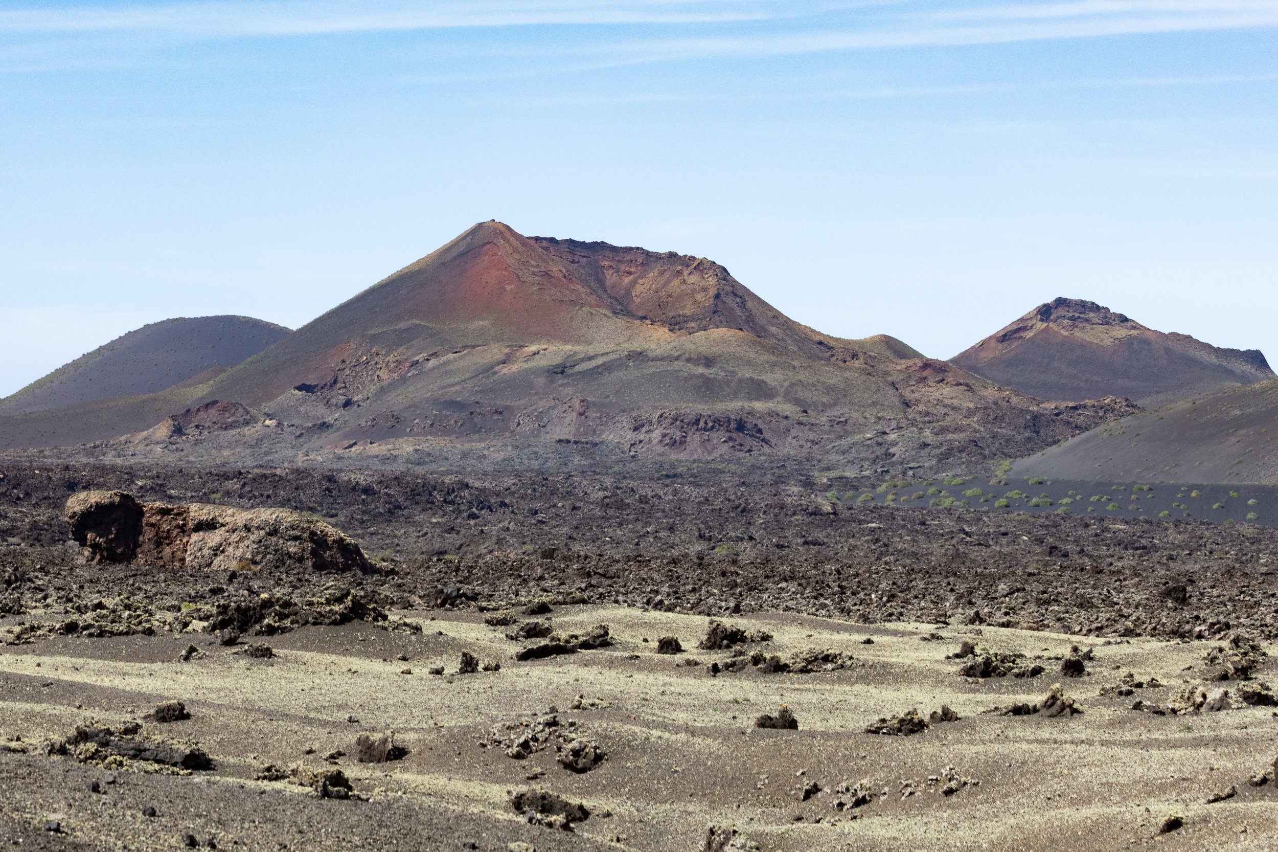 A volcanic landscape with dark, rocky terrain and volcanic mountains in the background under a blue sky.