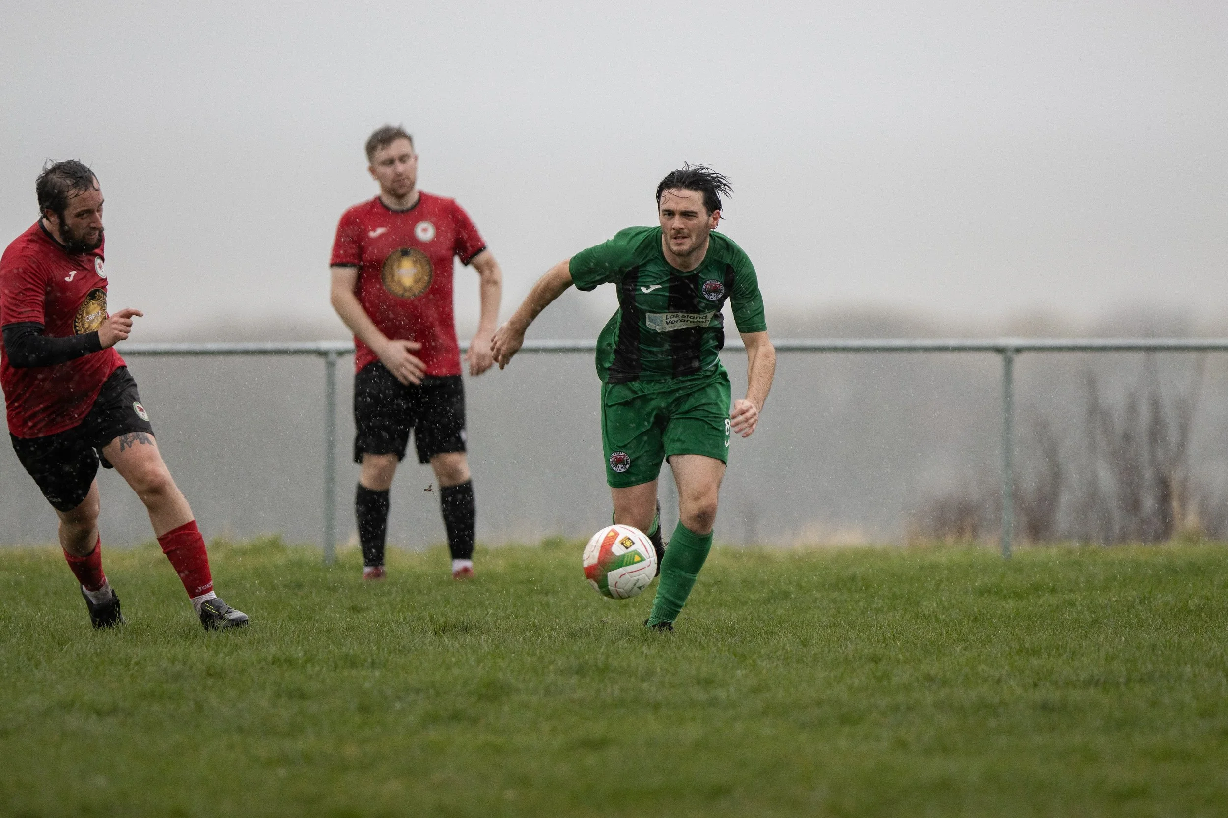 Soccer players in rain playing on wet field, one in green jersey dribbling ball, two in red jerseys running nearby.