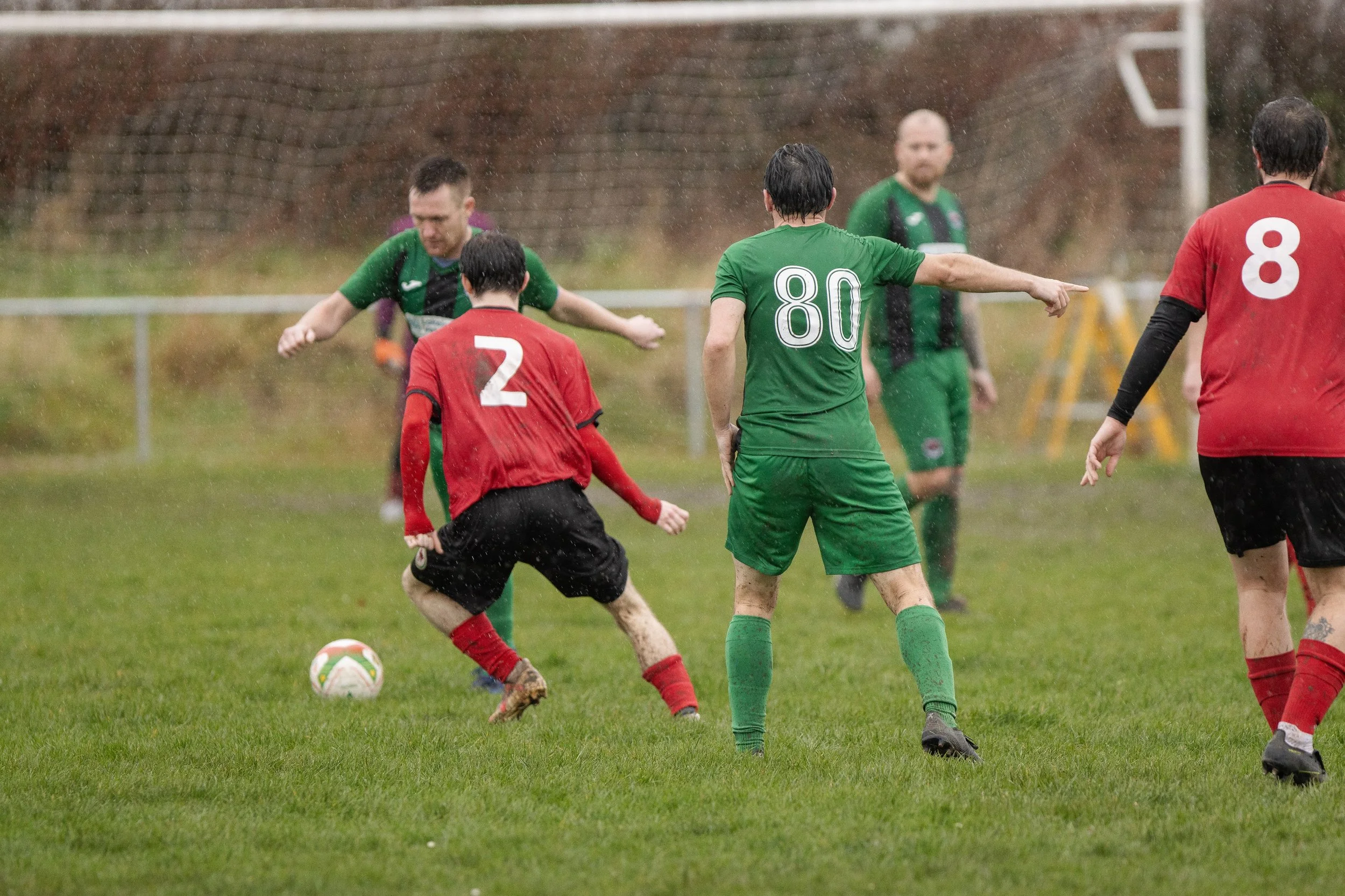 Soccer players on a rainy field, wearing red and green jerseys, competing for possession of the ball.