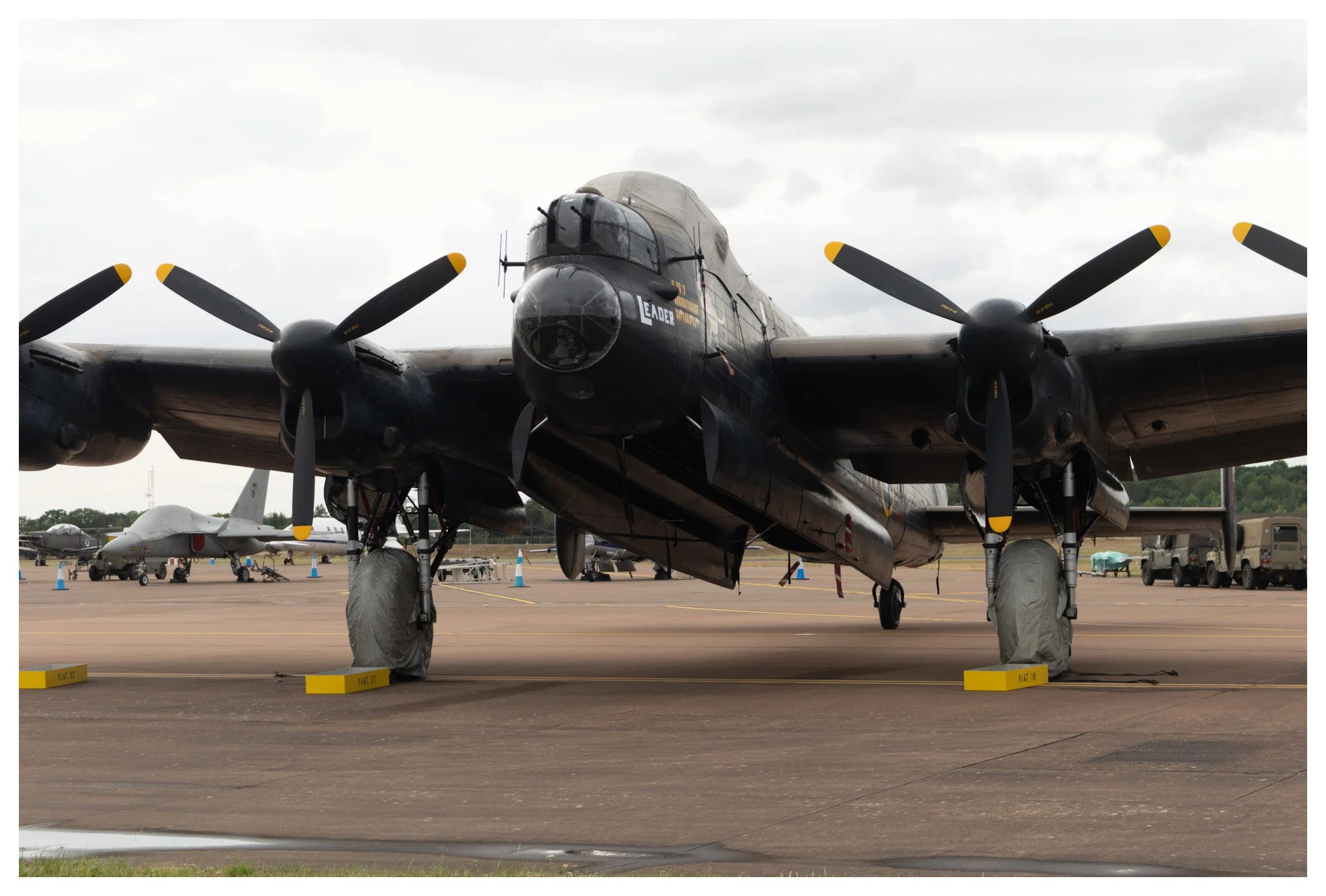Front view of a military bomber aircraft parked on an airfield, with several other military planes and vehicles in the background under gray cloudy skies.