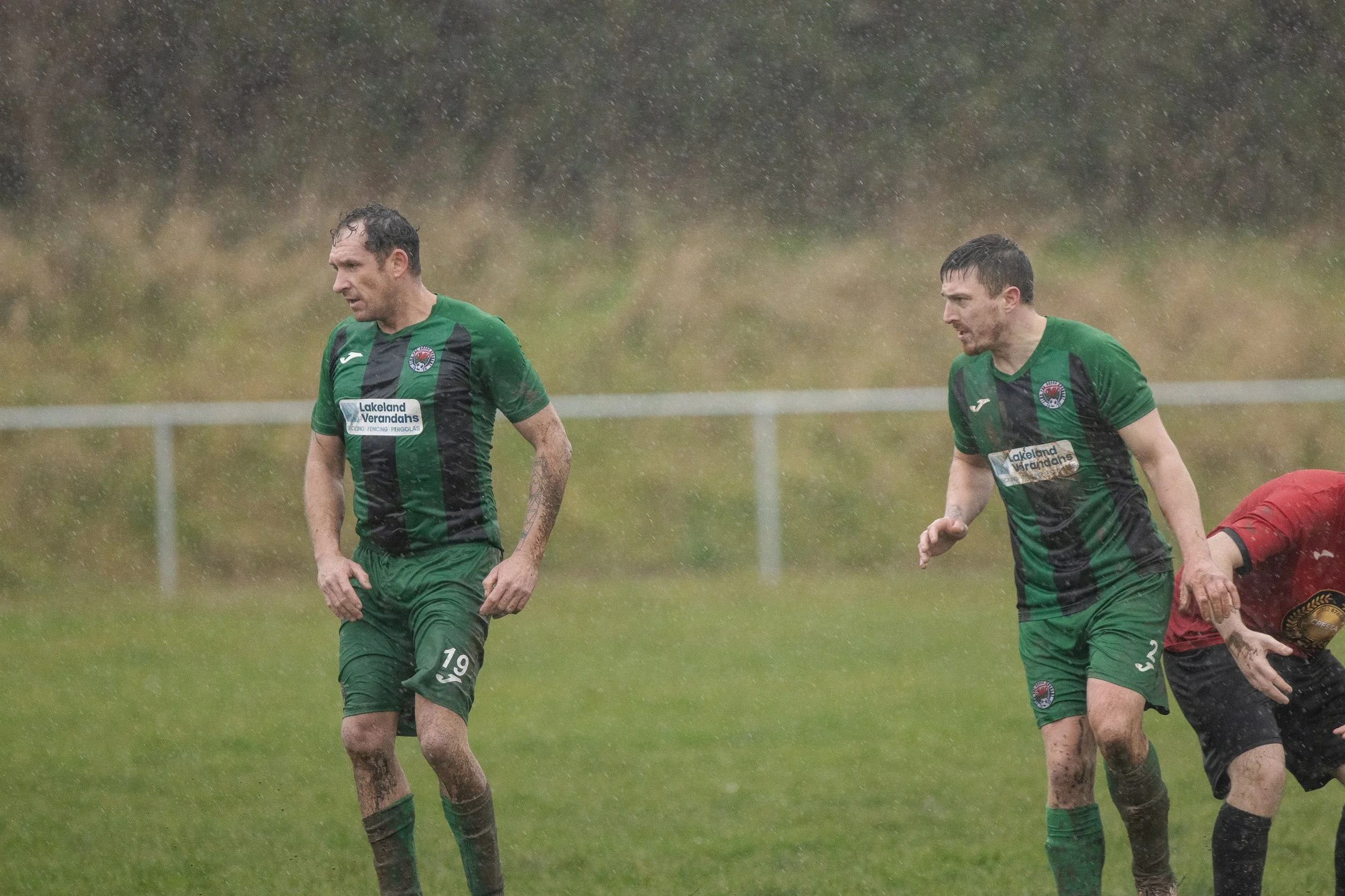 Two soccer players in green and black uniforms on a rainy field, with one player bent forward and the other standing, while a third person is partially visible to the right.