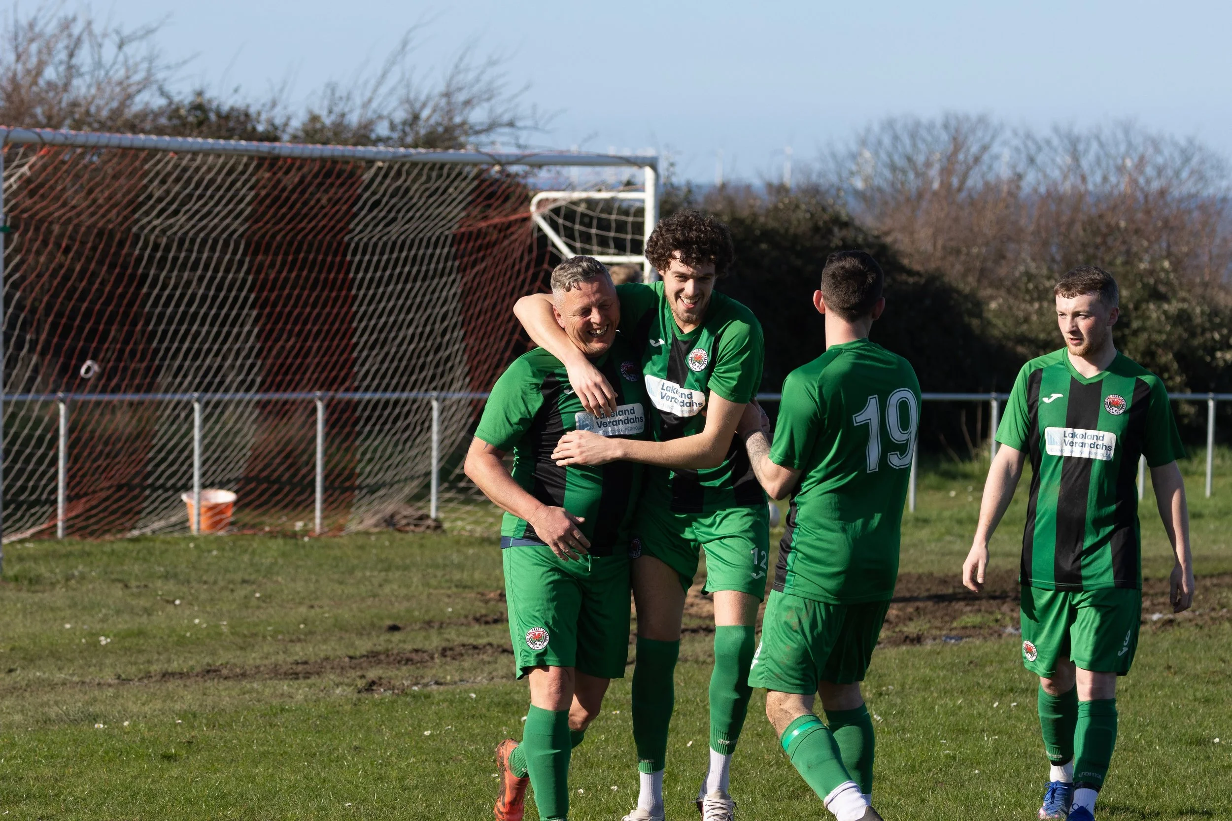 Soccer players in green uniforms celebrating on the field after scoring a goal, with a goalpost and net in the background.