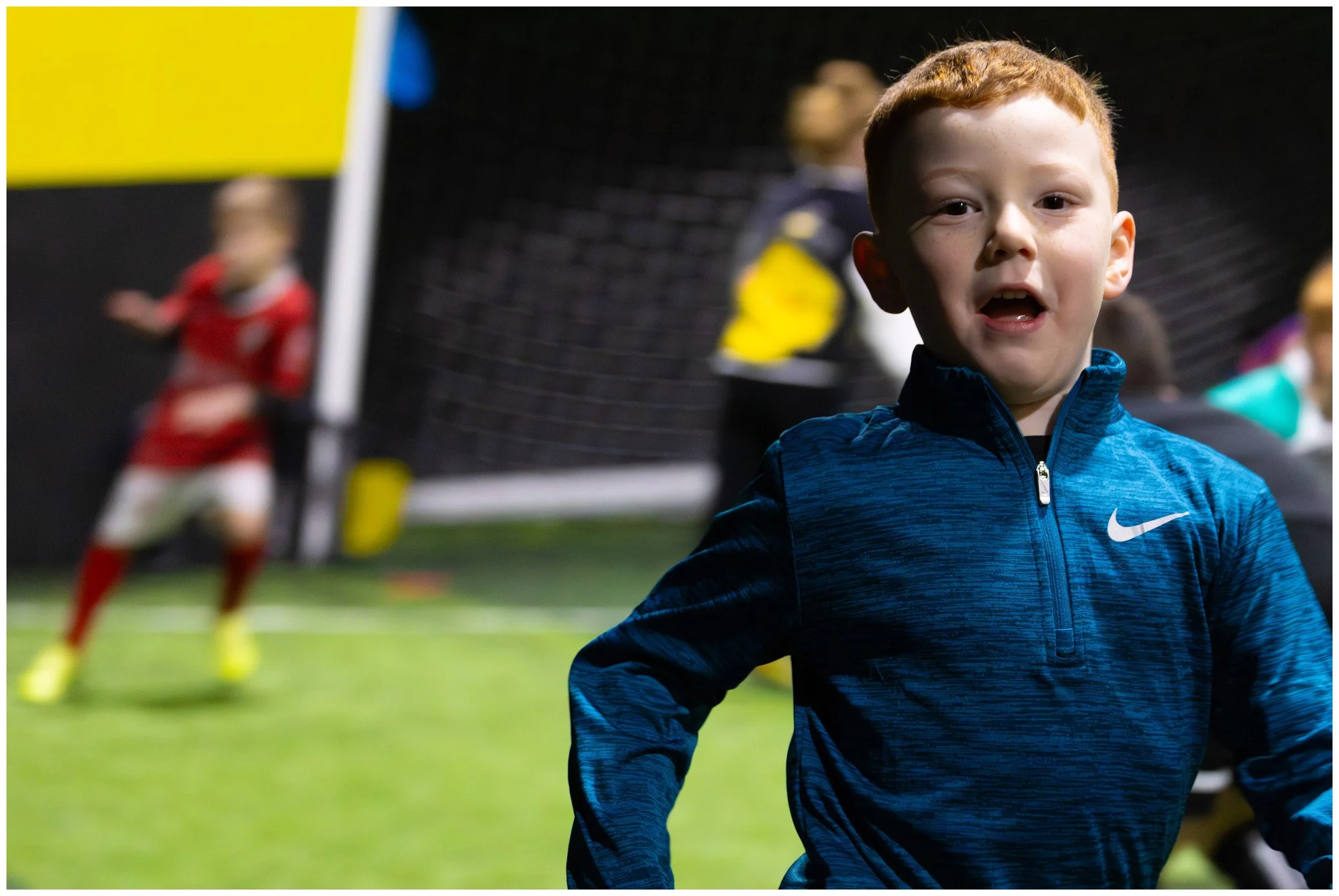 A young boy with red hair and freckles wearing a blue Nike sports jacket, standing on a soccer field during a game or practice, with other children and a goal net in the background.