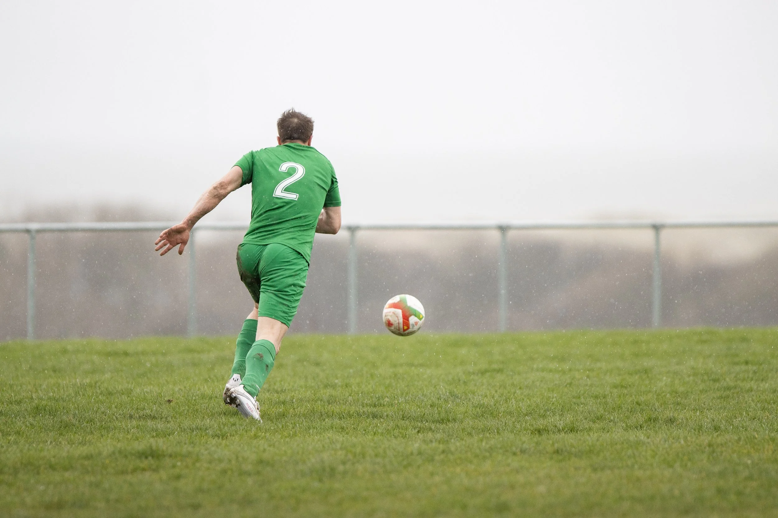 A soccer player in a green uniform with the number 2 on the back, running on a grassy field and about to kick a soccer ball under overcast skies.