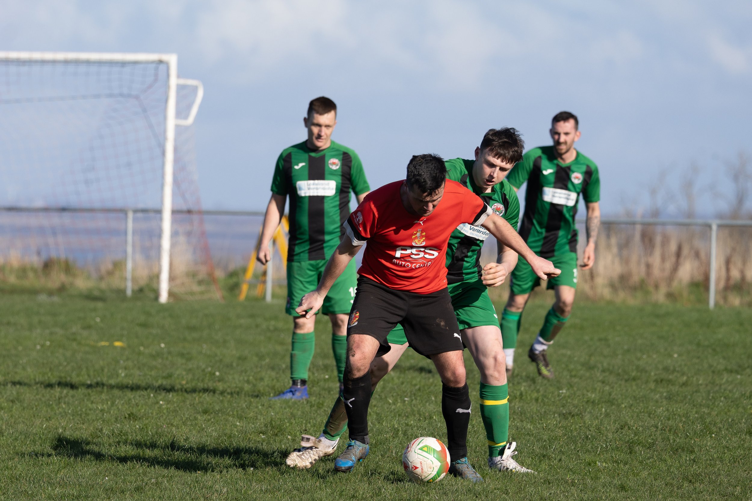 Four soccer players on a field, three in green jerseys and one in a red jersey, competing for the ball.