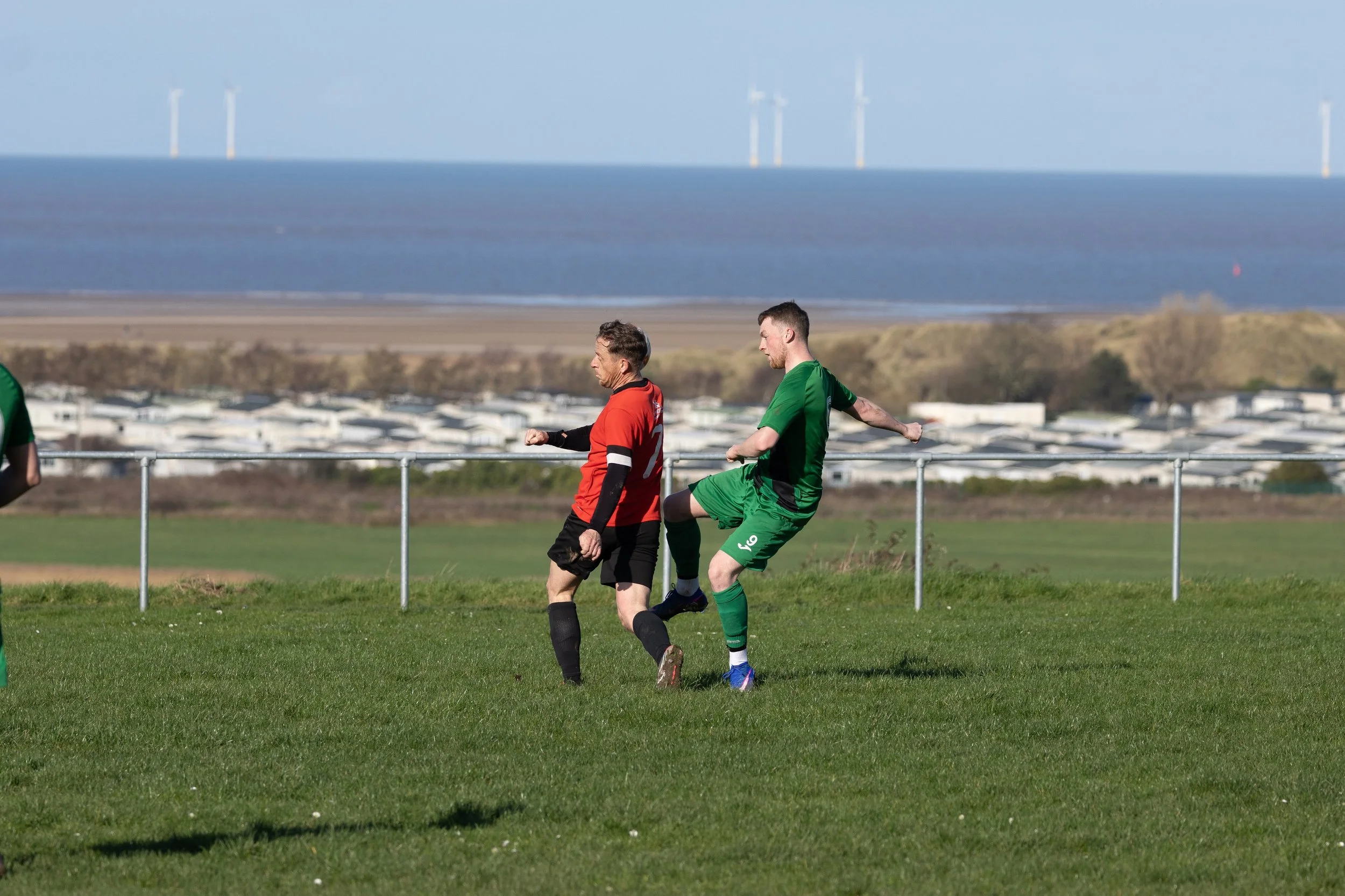 Two soccer players in green and red uniforms contesting for the ball during a match on a grassy field with a coastal background and wind turbines in the distance.