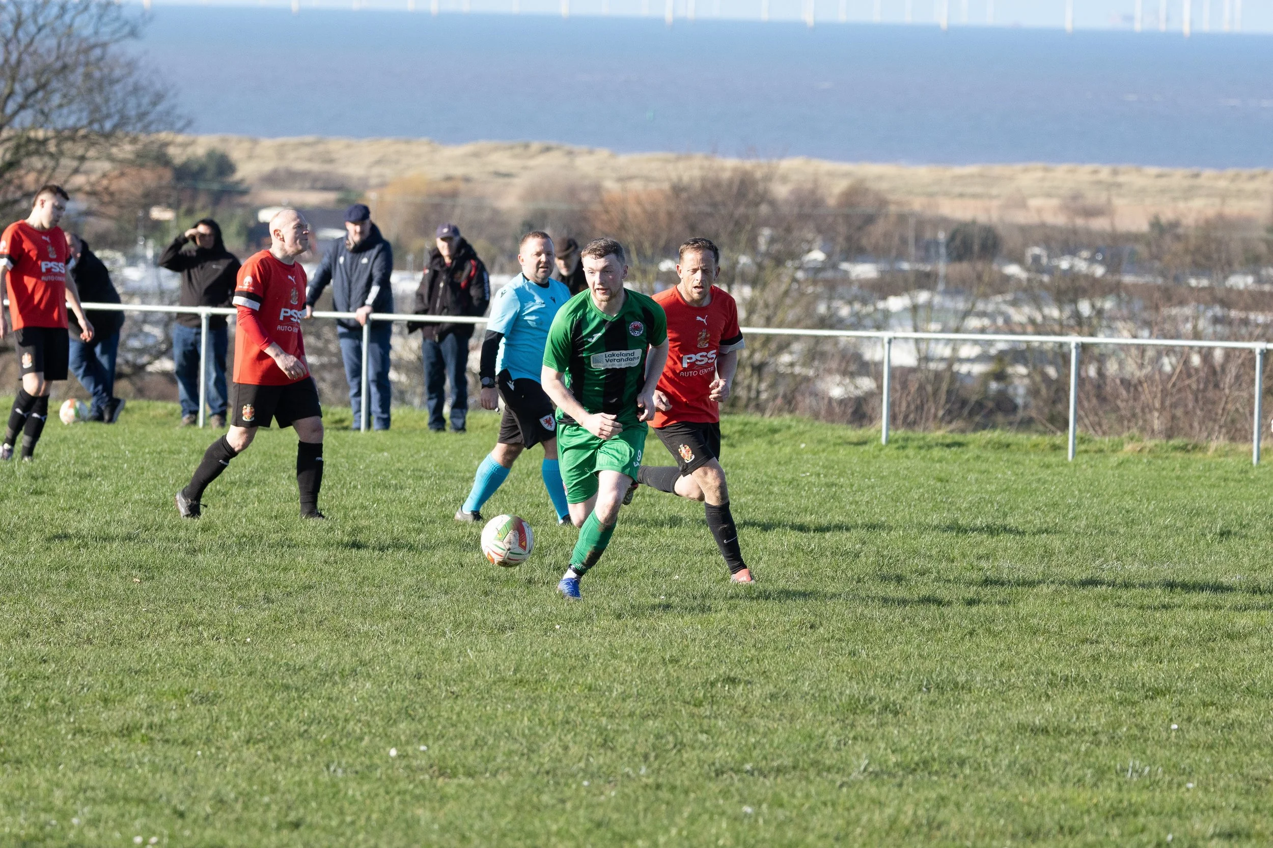 Soccer players in green and red jerseys playing on a grassy field with spectators watching, a scenic landscape with trees, houses, and a body of water in the background.