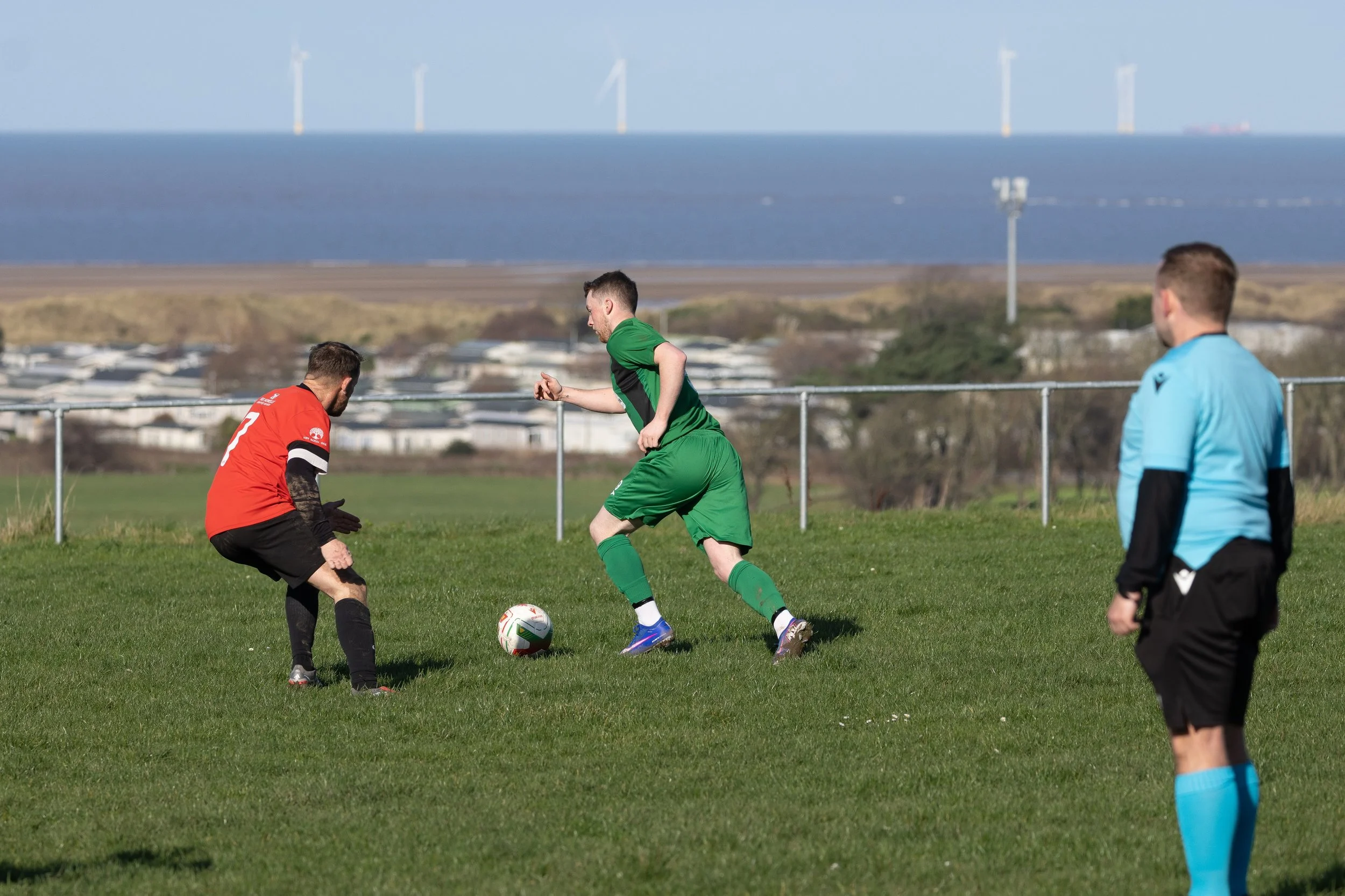 Soccer match on a grassy field with three players, one in a red jersey and black shorts, one in a green jersey and shorts, and an official in a light blue jersey and black shorts. The player in red appears to be guarding the ball while the player in 