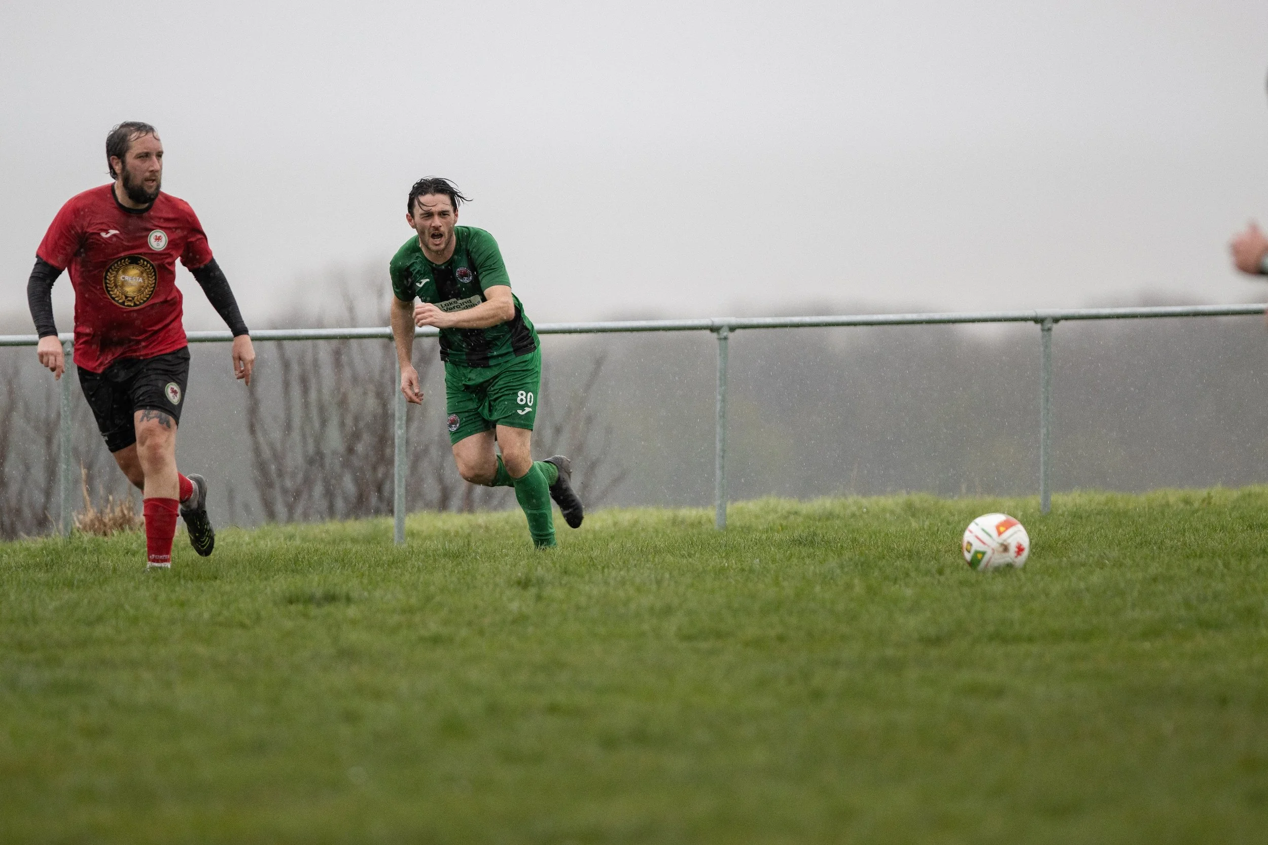 Two soccer players running on a grassy field in rainy weather, chasing the soccer ball, with gray overcast sky in the background.