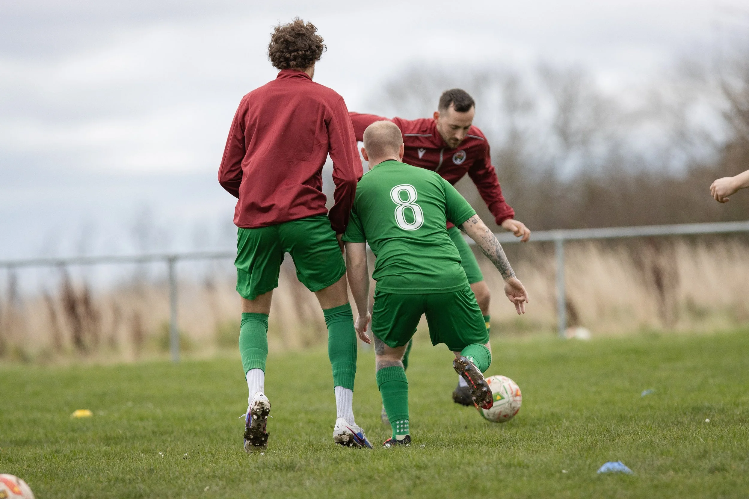 Three soccer players on the field during a game, two in green uniforms and one in a red jacket, competing for ball possession amidst cloudy weather.