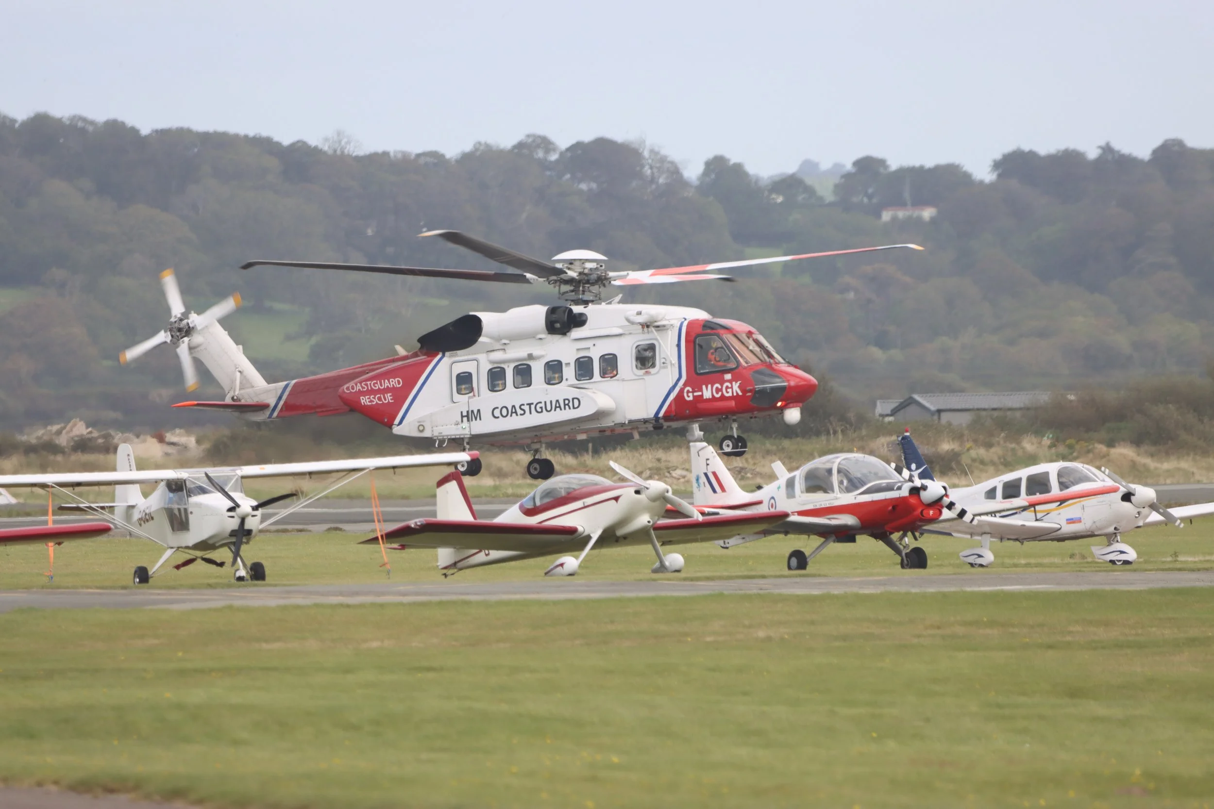 Multiple aircraft, including a large rescue helicopter and smaller planes, parked on an airfield with a grassy landscape and trees in the background.