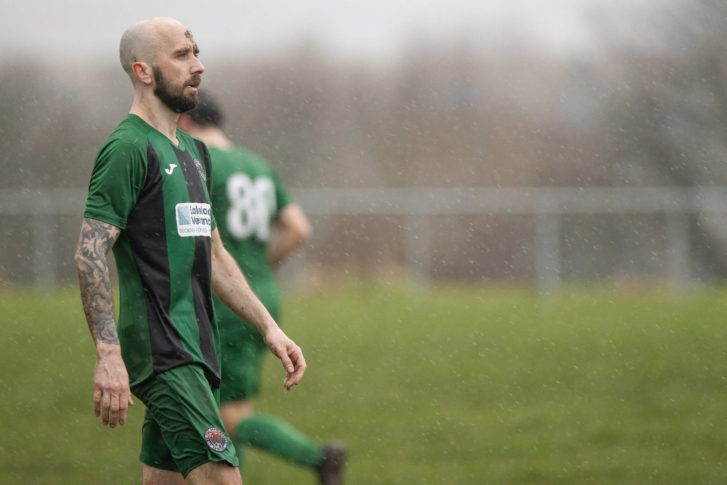 A male soccer player with a beard, tattoos on his arms, and a bald head, wearing a black and green uniform, walking on the field in the rain.
