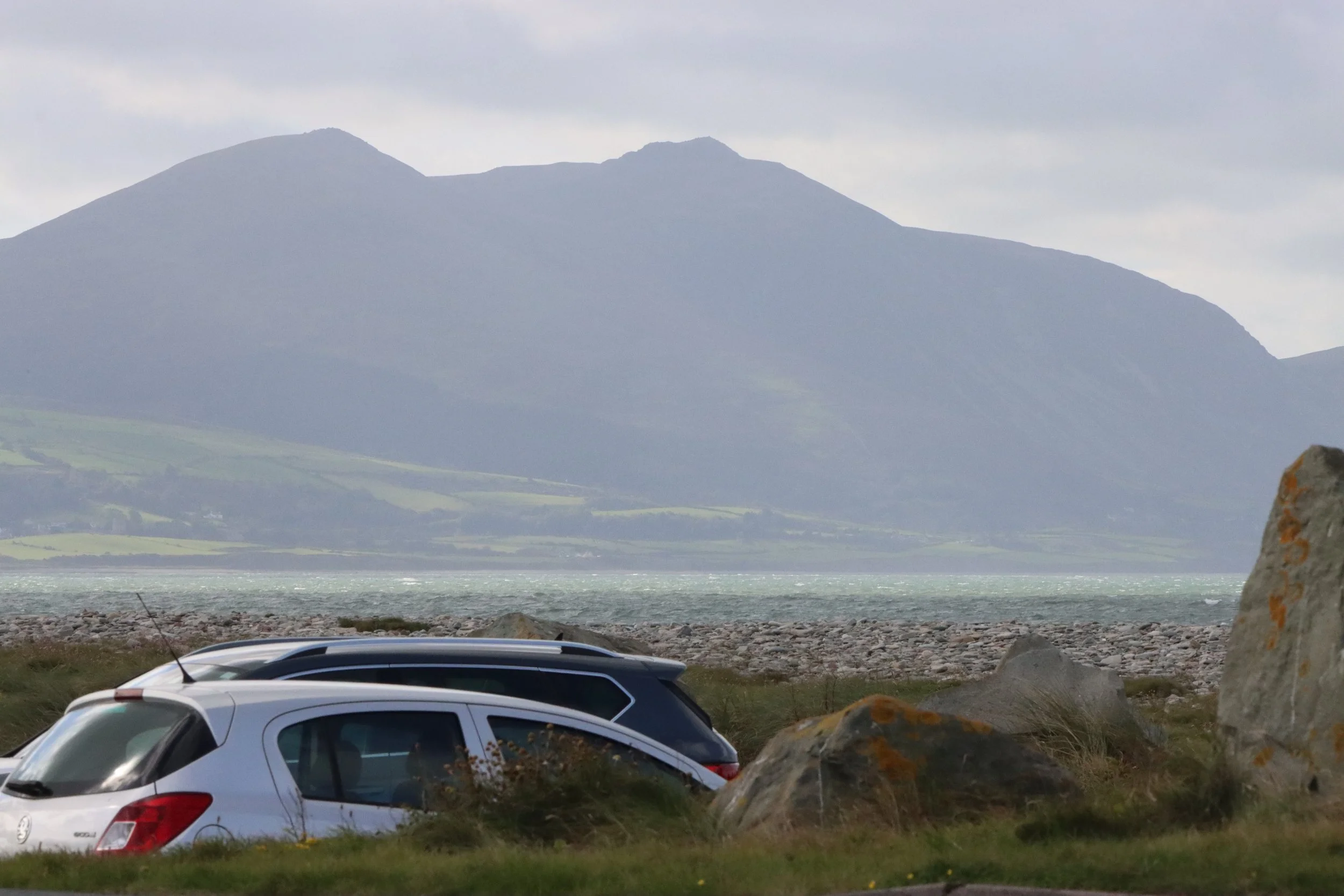 White cars parked on a grassy and rocky area with large rocks in the foreground, with a body of water and mountains in the background under a cloudy sky.