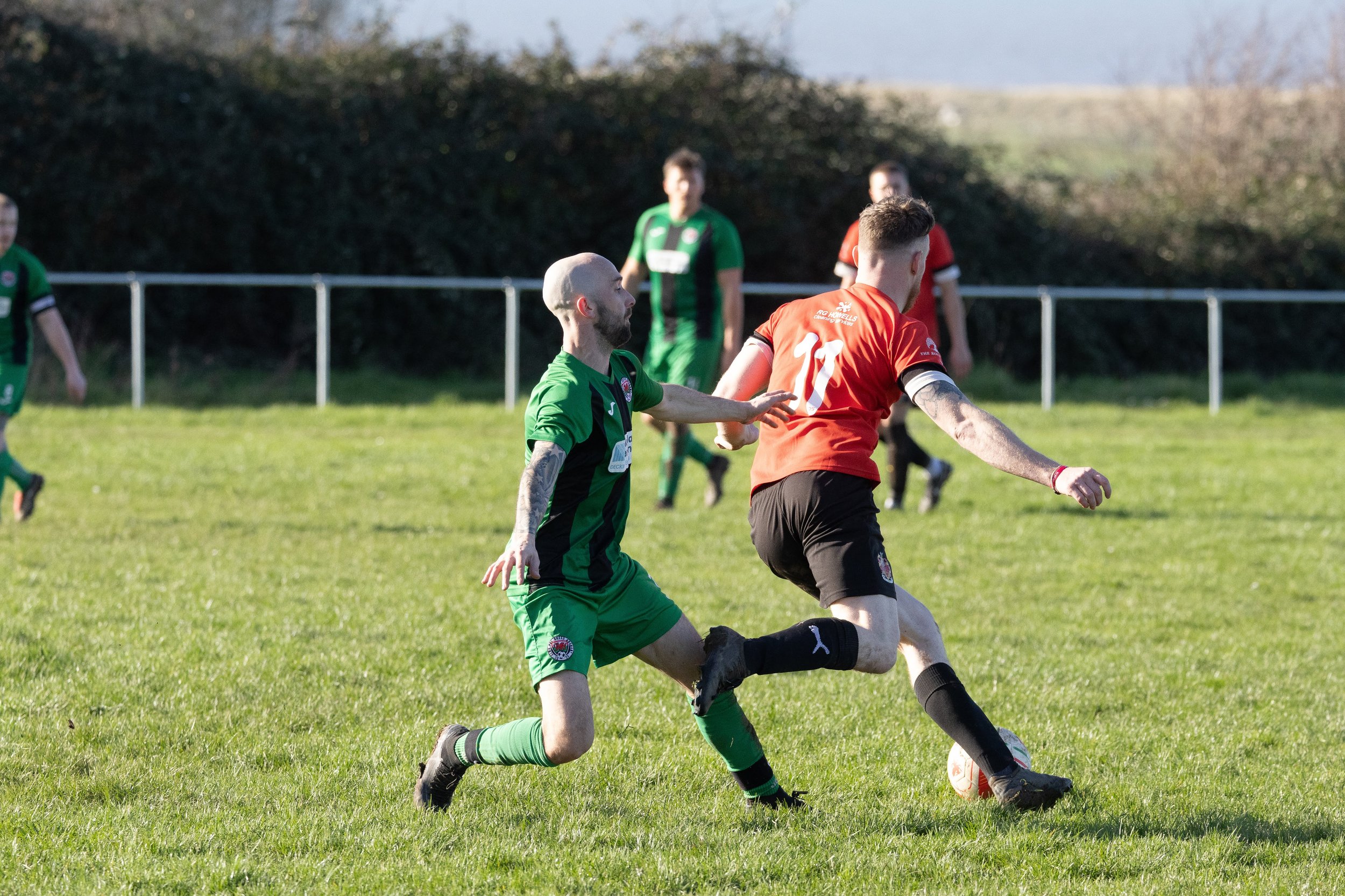 Two soccer players, one in a green and black uniform and the other in a red and black uniform, competing for the ball on a grassy field with three players in the background.