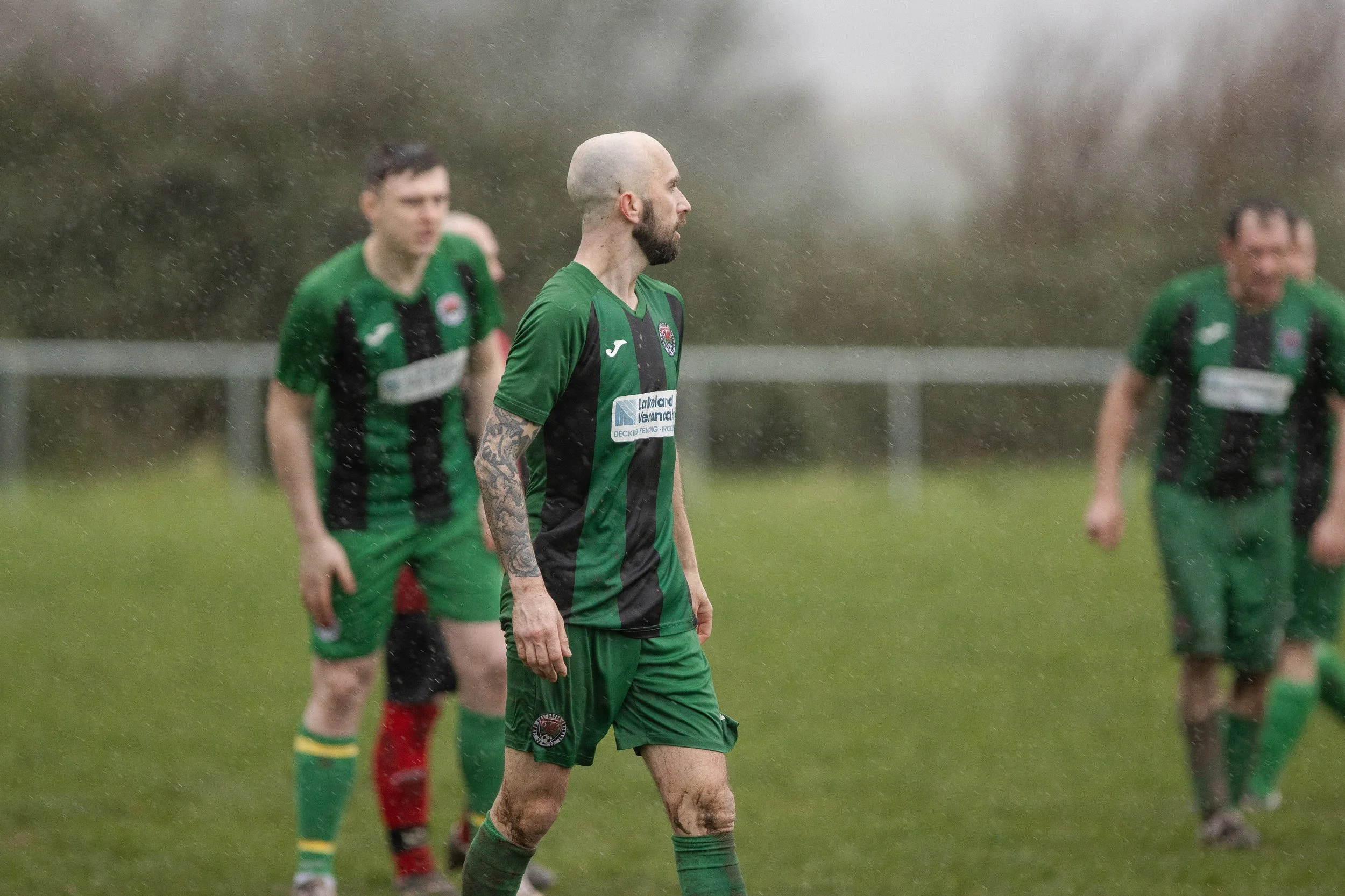 Soccer players in green and black uniforms standing on a muddy, rainy field during a game.