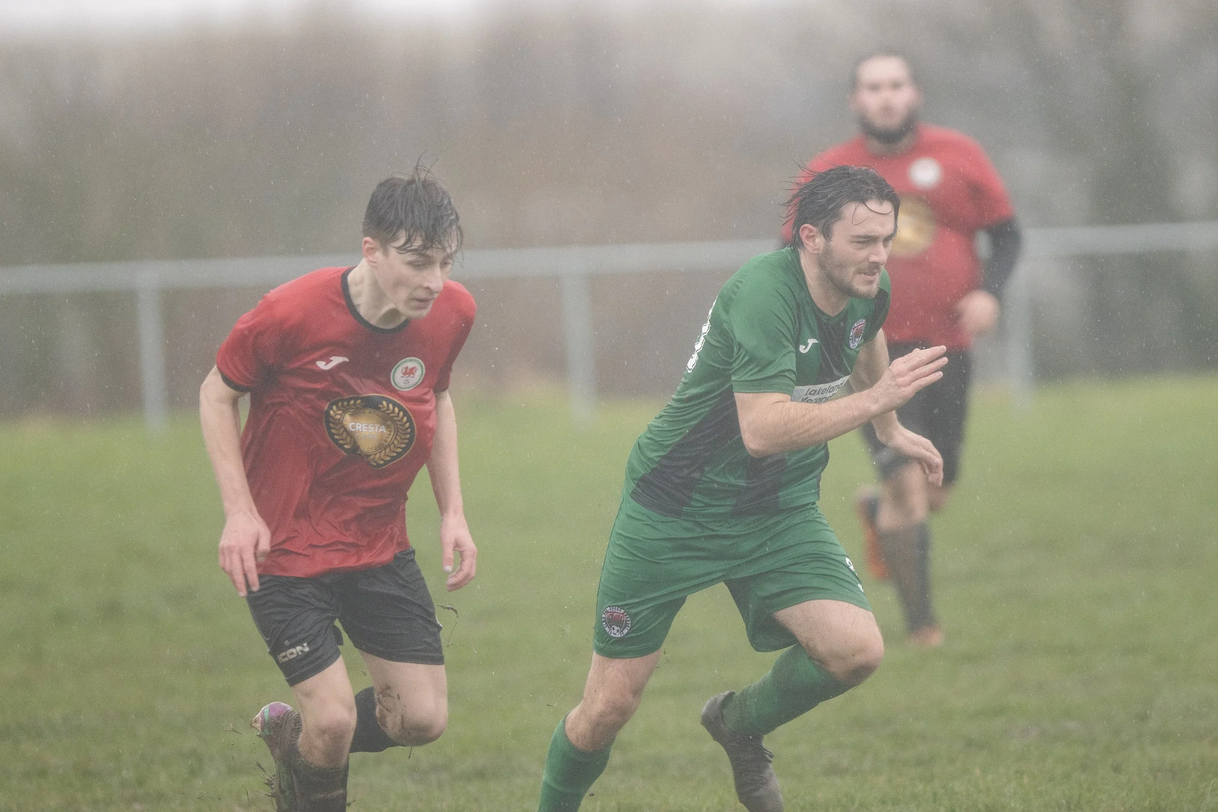 Two soccer players running in the rain on a grassy field, one in a red jersey and the other in a green jersey, with another player in red in the background.