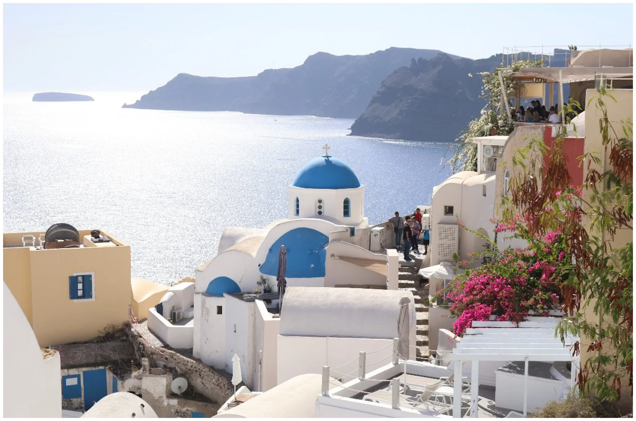 A scenic view of white buildings with blue domes on a cliffside overlooking the ocean in Santorini, Greece. Some buildings have outdoor terraces with people, and pink flowering bushes are in the foreground.