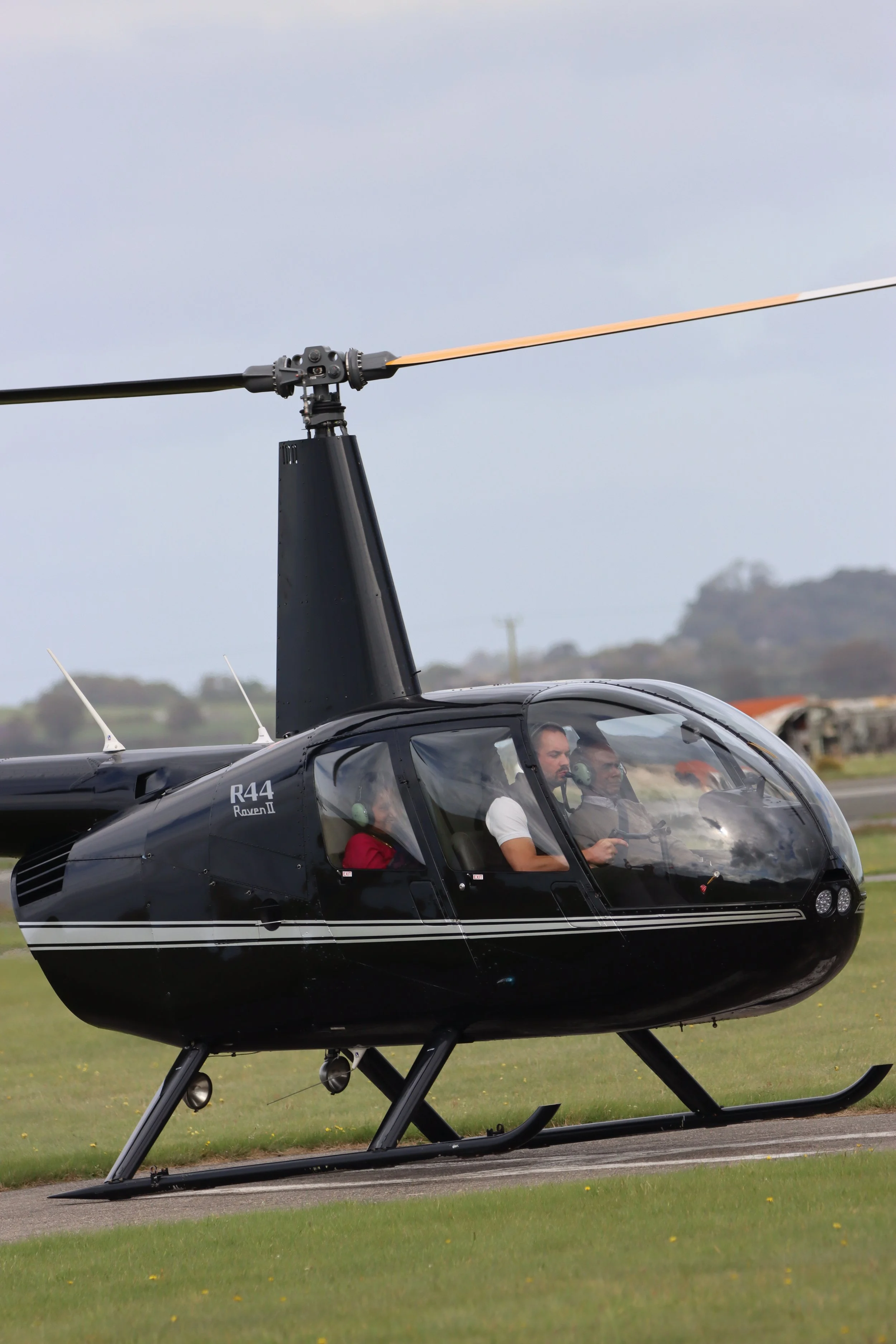 Black Robinson R44 helicopter on the ground with its rotor blades spinning, with three people inside, two in the front and one in the back, on a grassy area during daytime.