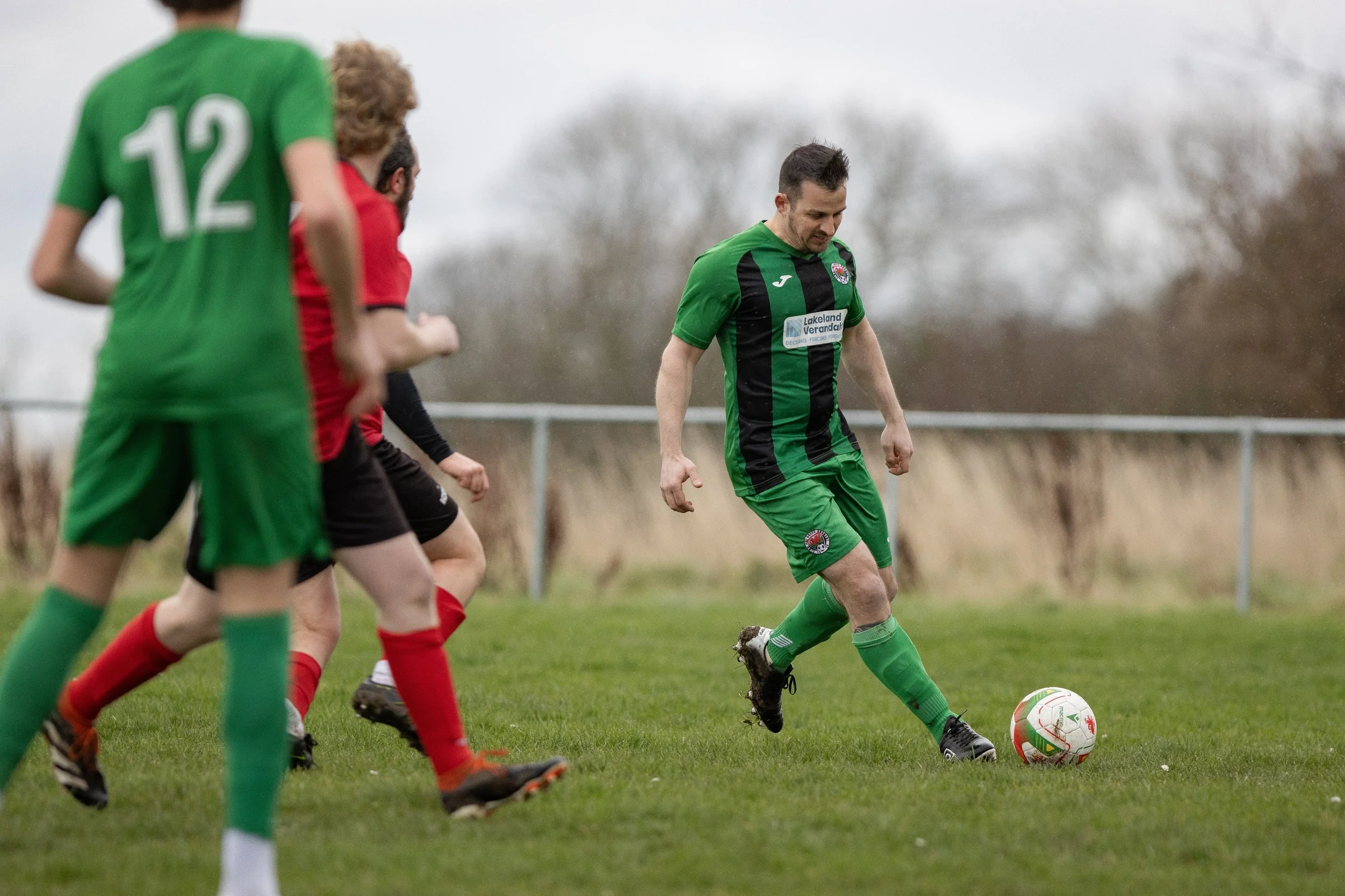 A soccer player in a green and black uniform is kicking a ball during a match, with other players in red and black uniforms running nearby on the field.