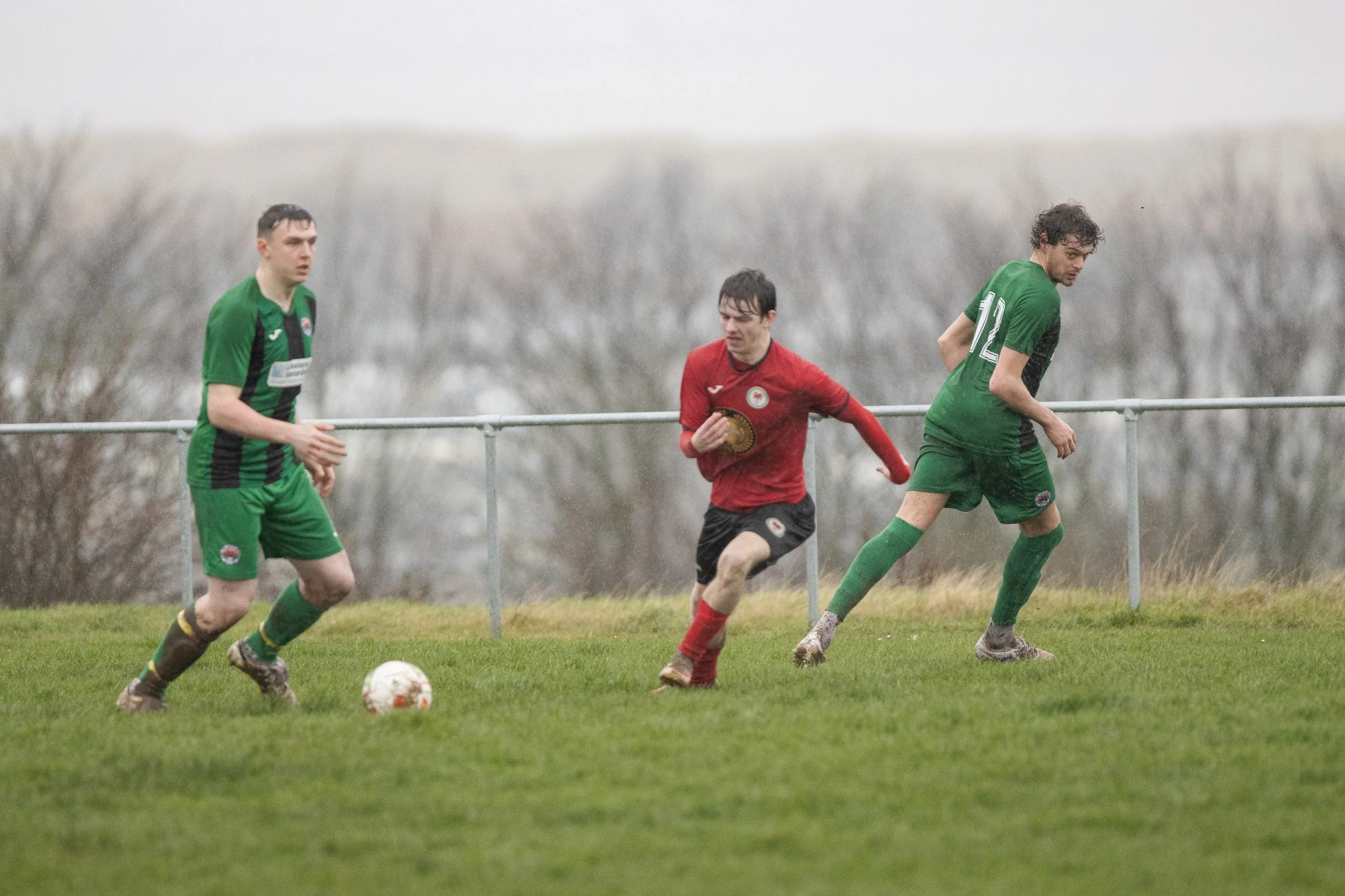 Three soccer players on a grassy field, two in green jerseys and one in red, during a rainy game, with a background of leafless trees and overcast sky.