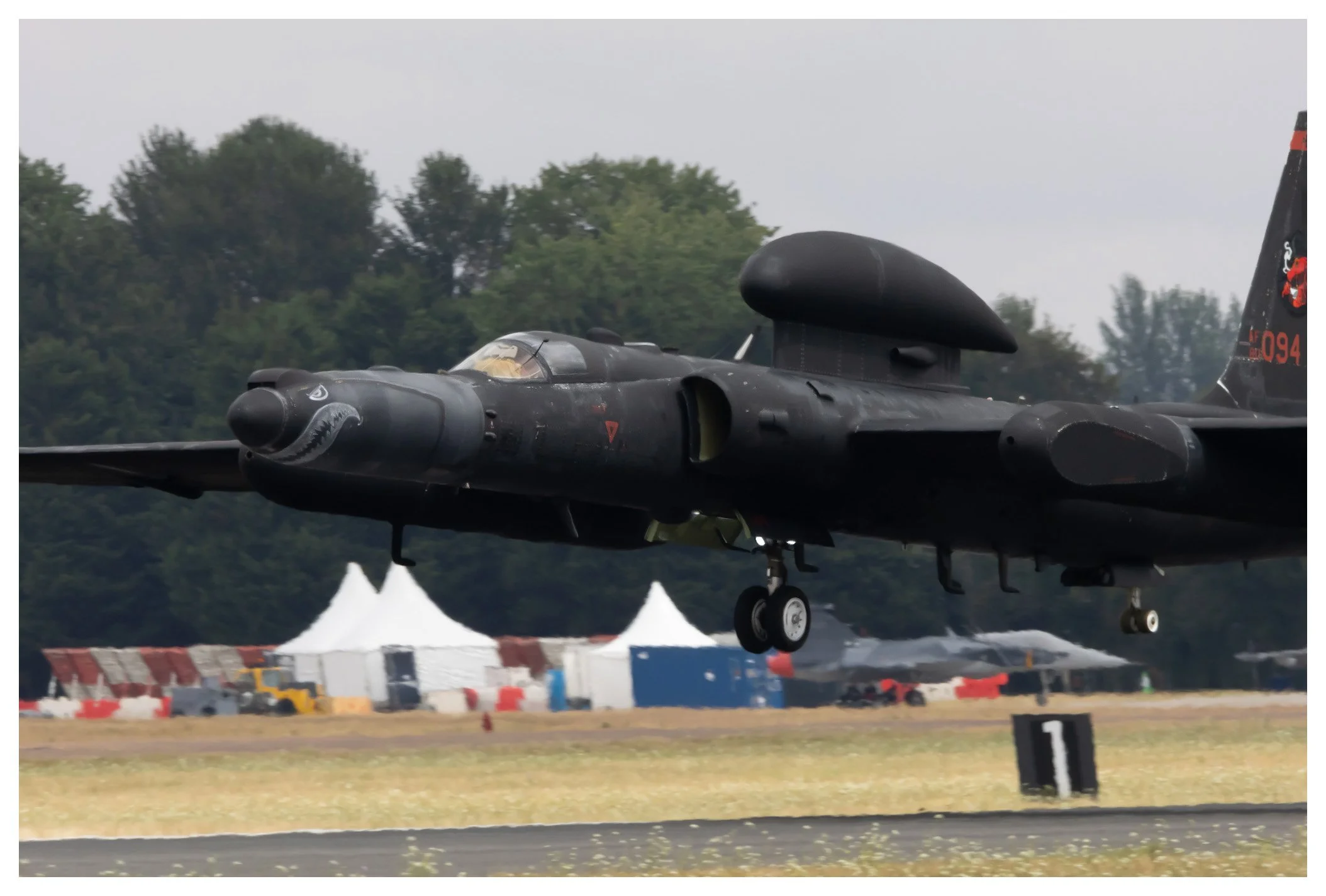 A black military jet plane with shark mouth nose art taking off from a runway, with tents and trees in the background.