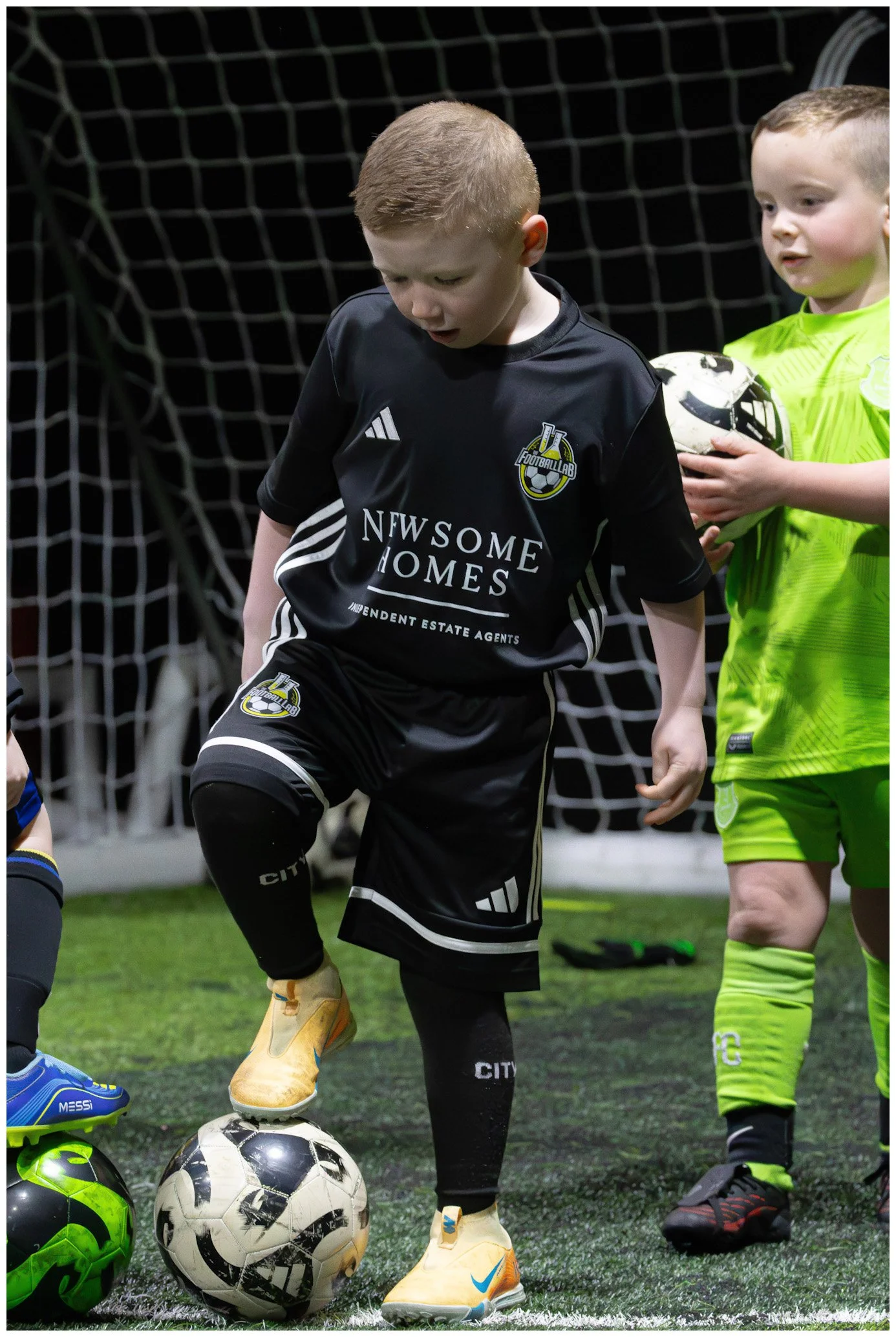 Young boys participating in indoor soccer, wearing sports uniforms and additional soccer balls on the artificial turf field.
