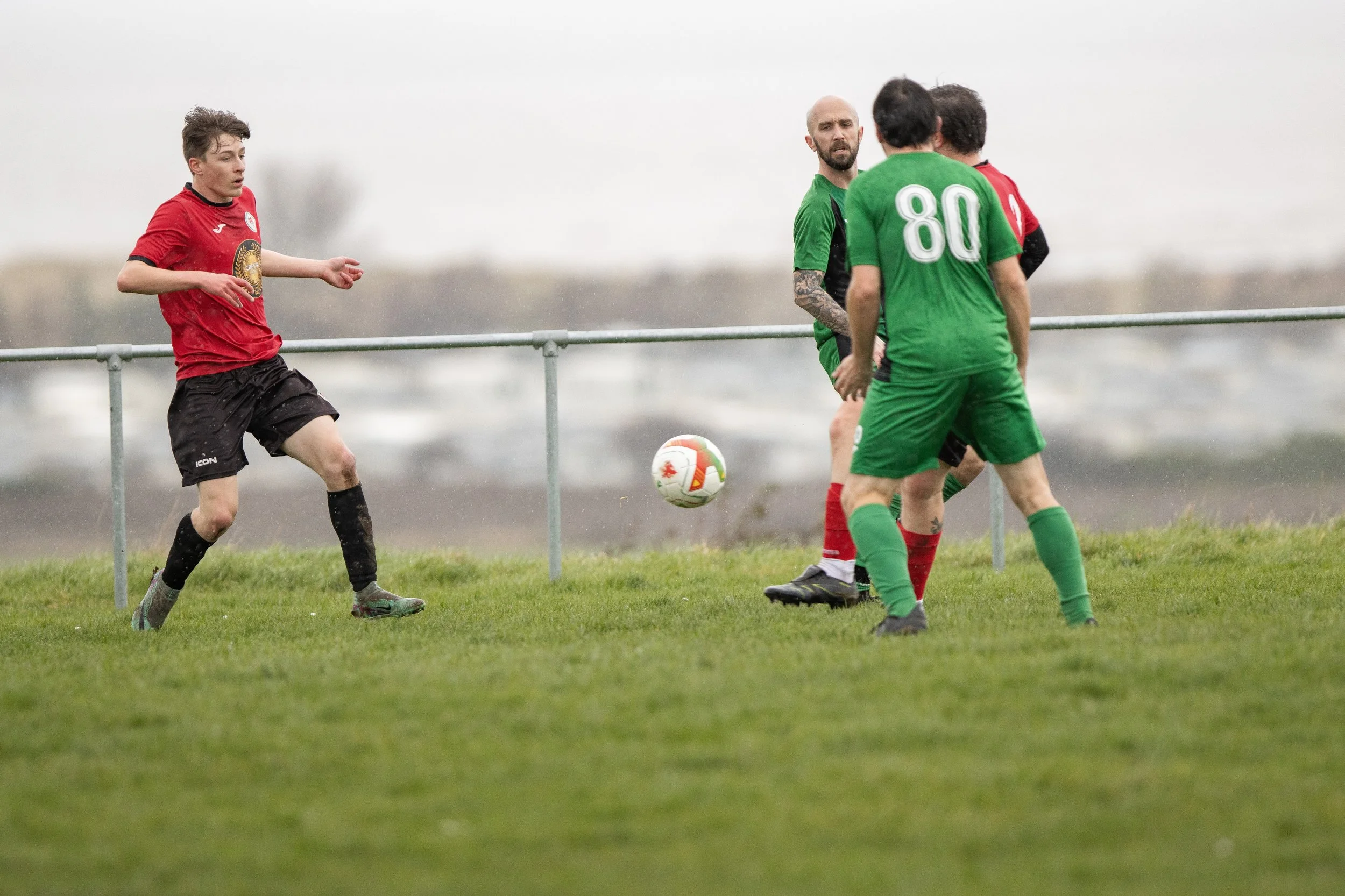 Soccer players on a grassy field, two in red and two in green, near a metal railing on a cloudy day.