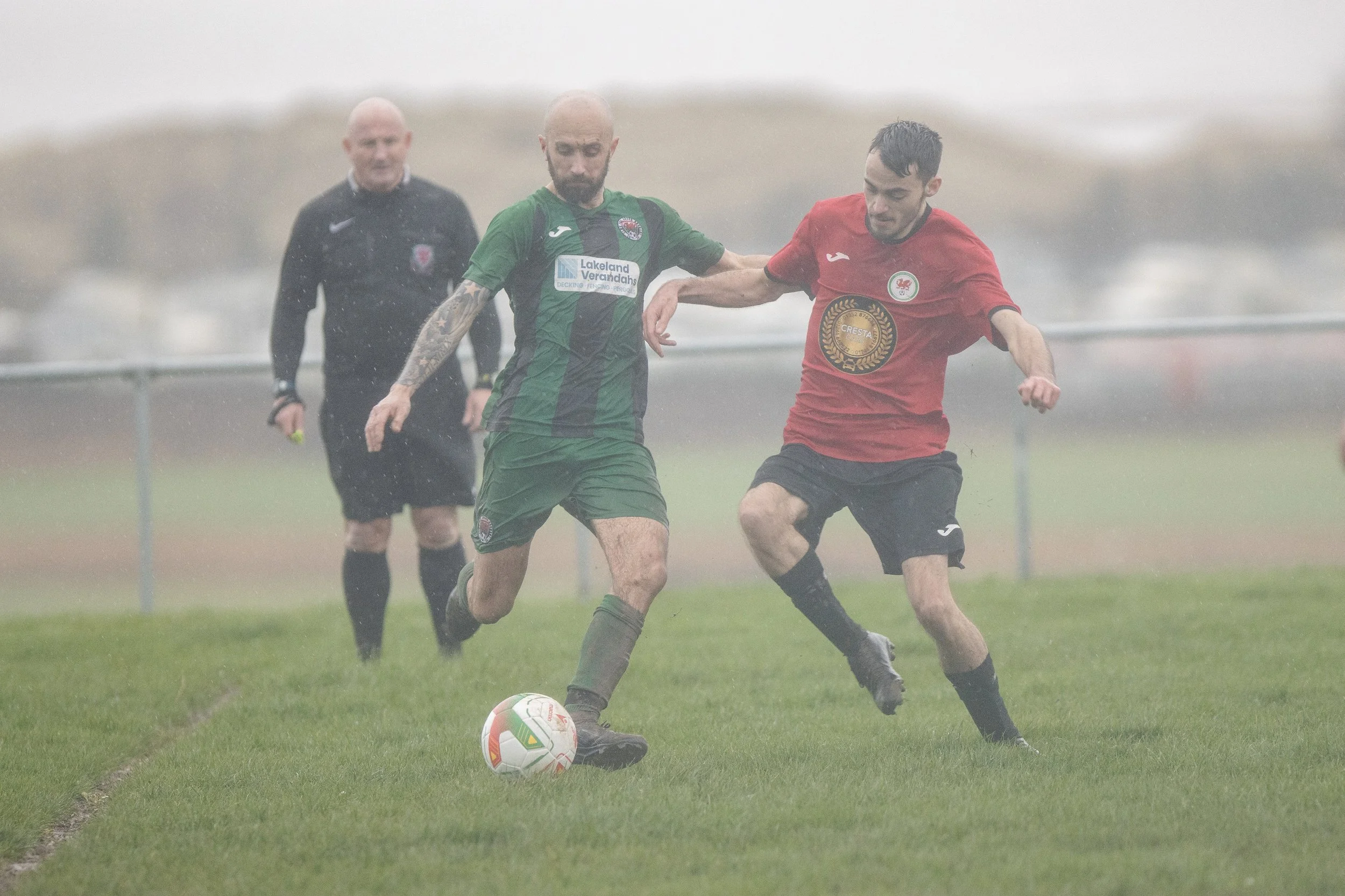 Two soccer players with tattoos and dark hair compete for possession of the ball on a rainy field, with a referee in black observing in the background.