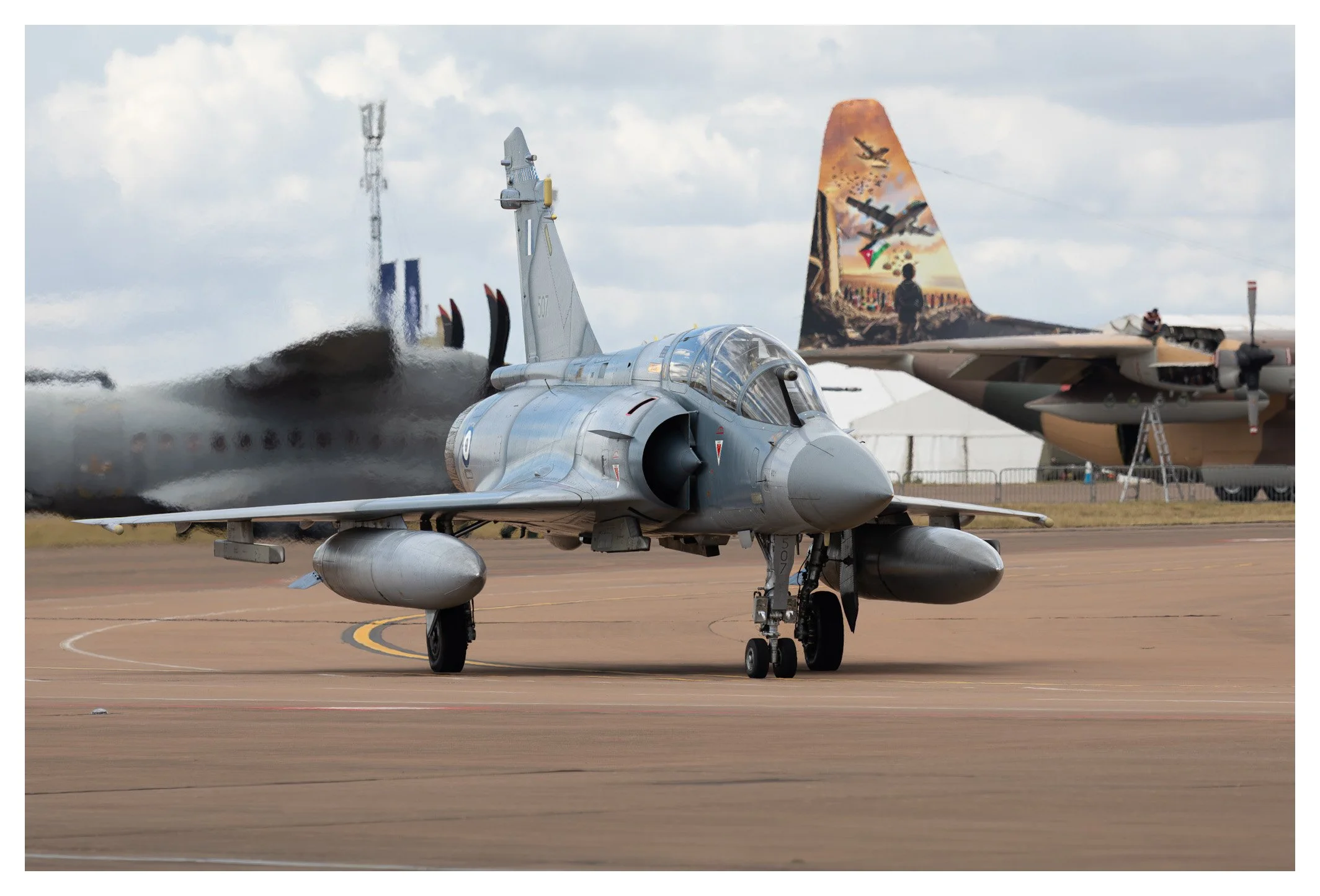 A military fighter jet taxiing on the runway with another aircraft in the background and a mural on its tail fin. The fighter jet has a sleek, gray body with missiles under its wings.