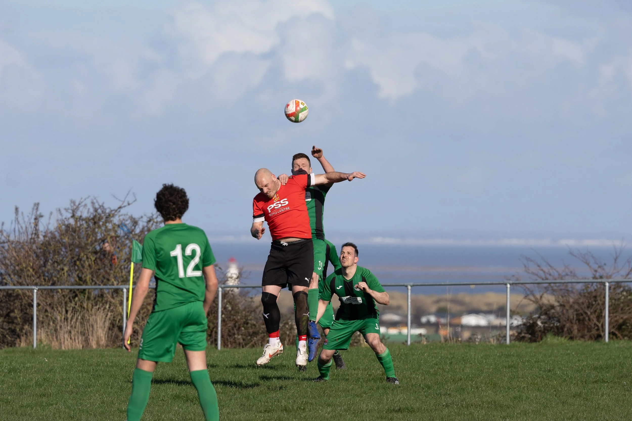 Four soccer players in green and red uniforms competing for a ball in the air during a game on a grassy field.