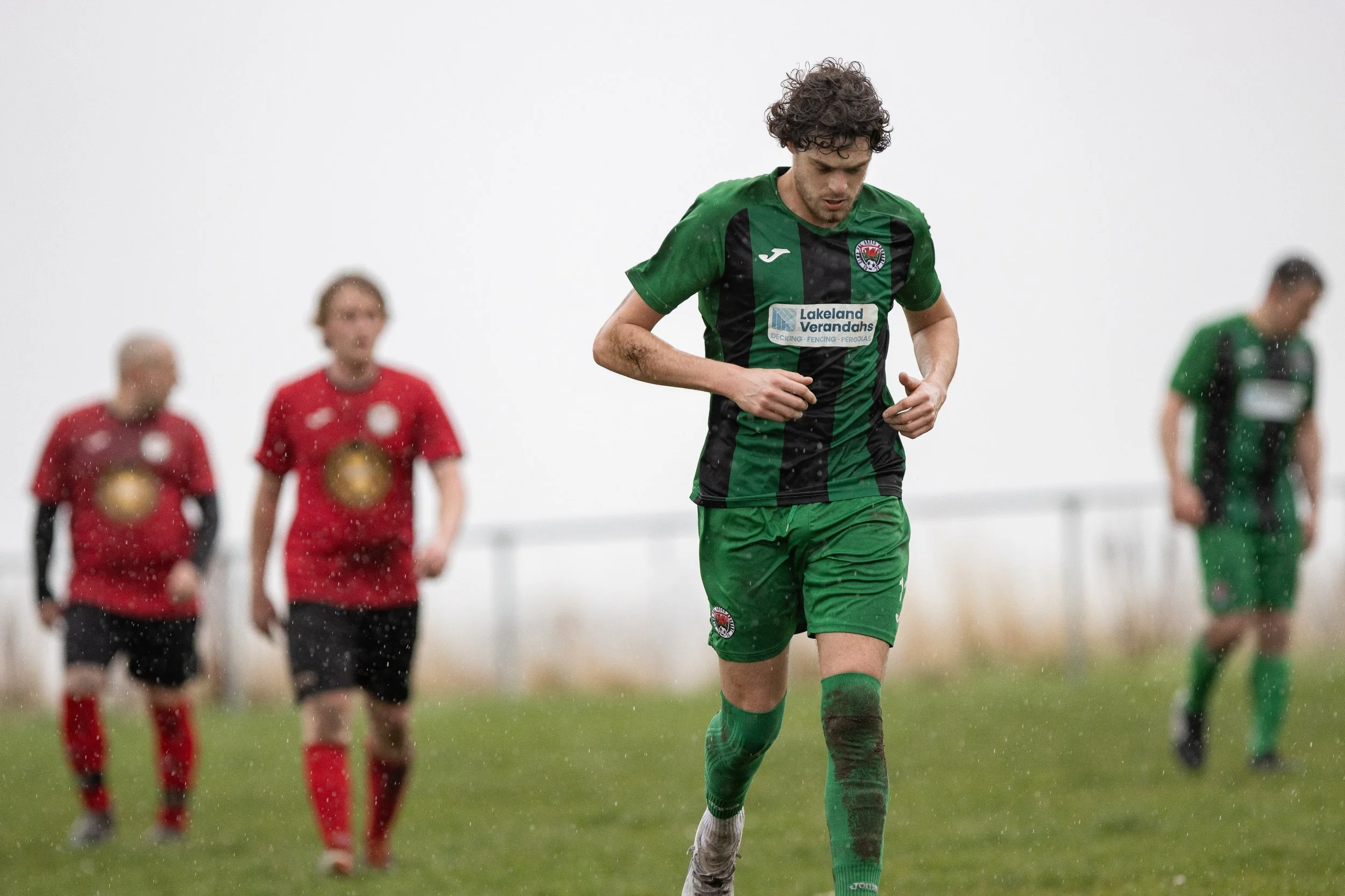 A soccer player in a green and black uniform running on a rainy field with three other players in red and one in green visible in the background.