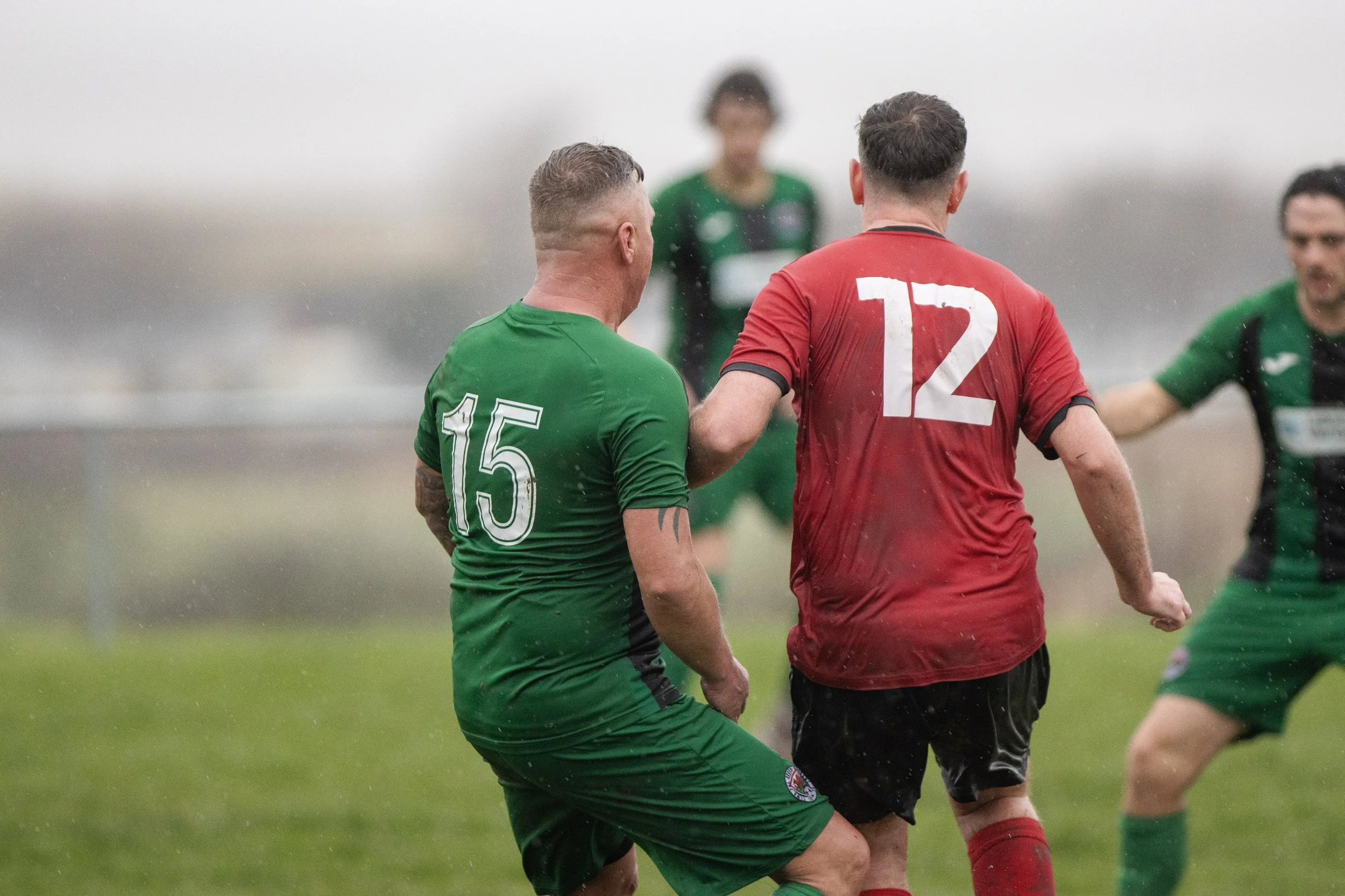 Soccer players in green and red uniforms during match in rainy weather, with one player holding another by the shoulder.