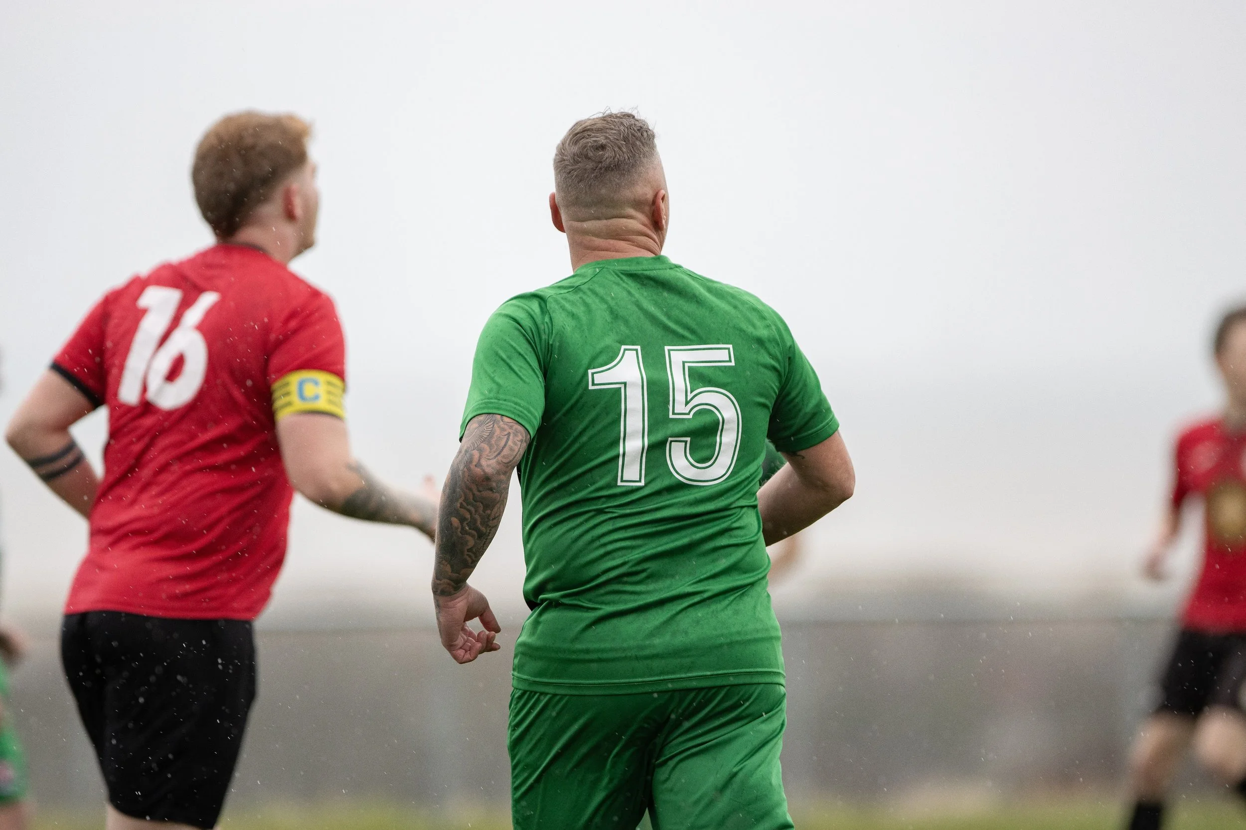 Soccer players on the field, one in a green jersey with the number 15, and others in red jerseys, during a match in rainy weather.