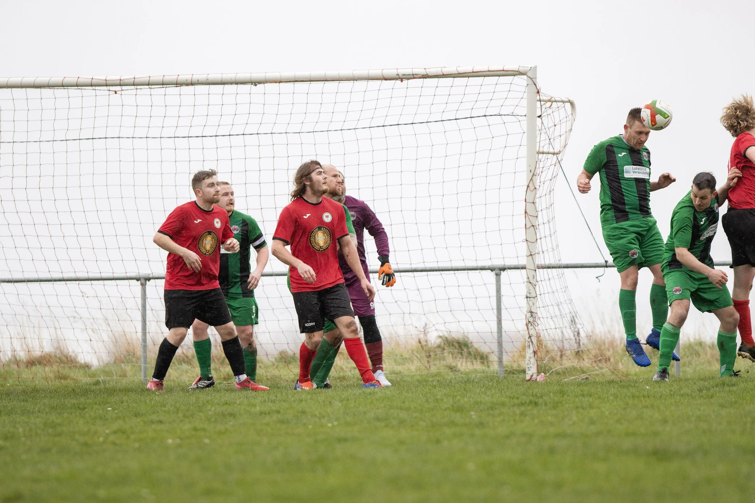 Soccer players in red and green uniforms compete near the goal on a grassy field with overcast sky.