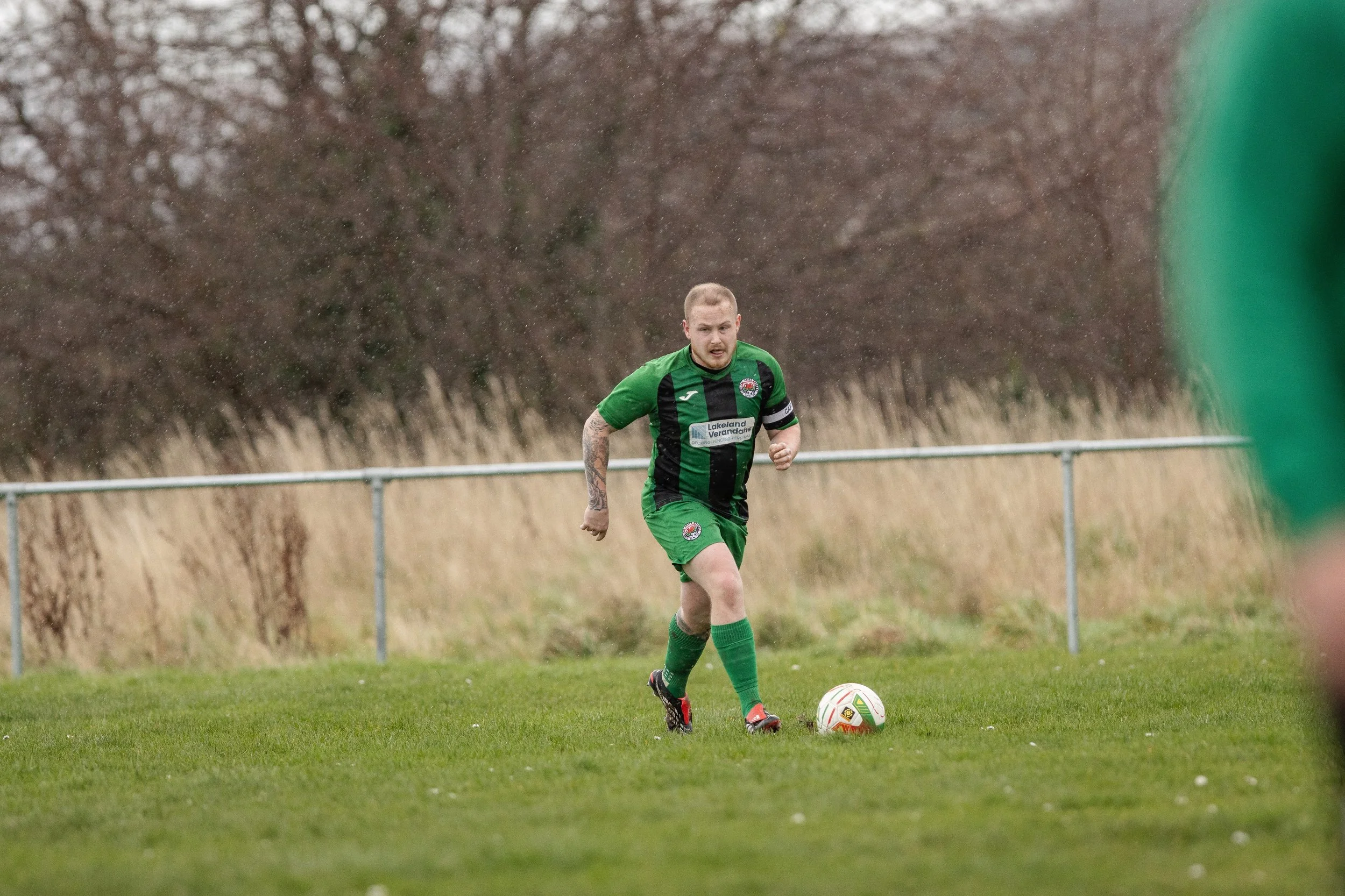 A male soccer player in a green and black uniform is running on a grass field with a soccer ball at his feet. The background shows brown trees and a metal fence, suggesting an outdoor field on a cloudy day.