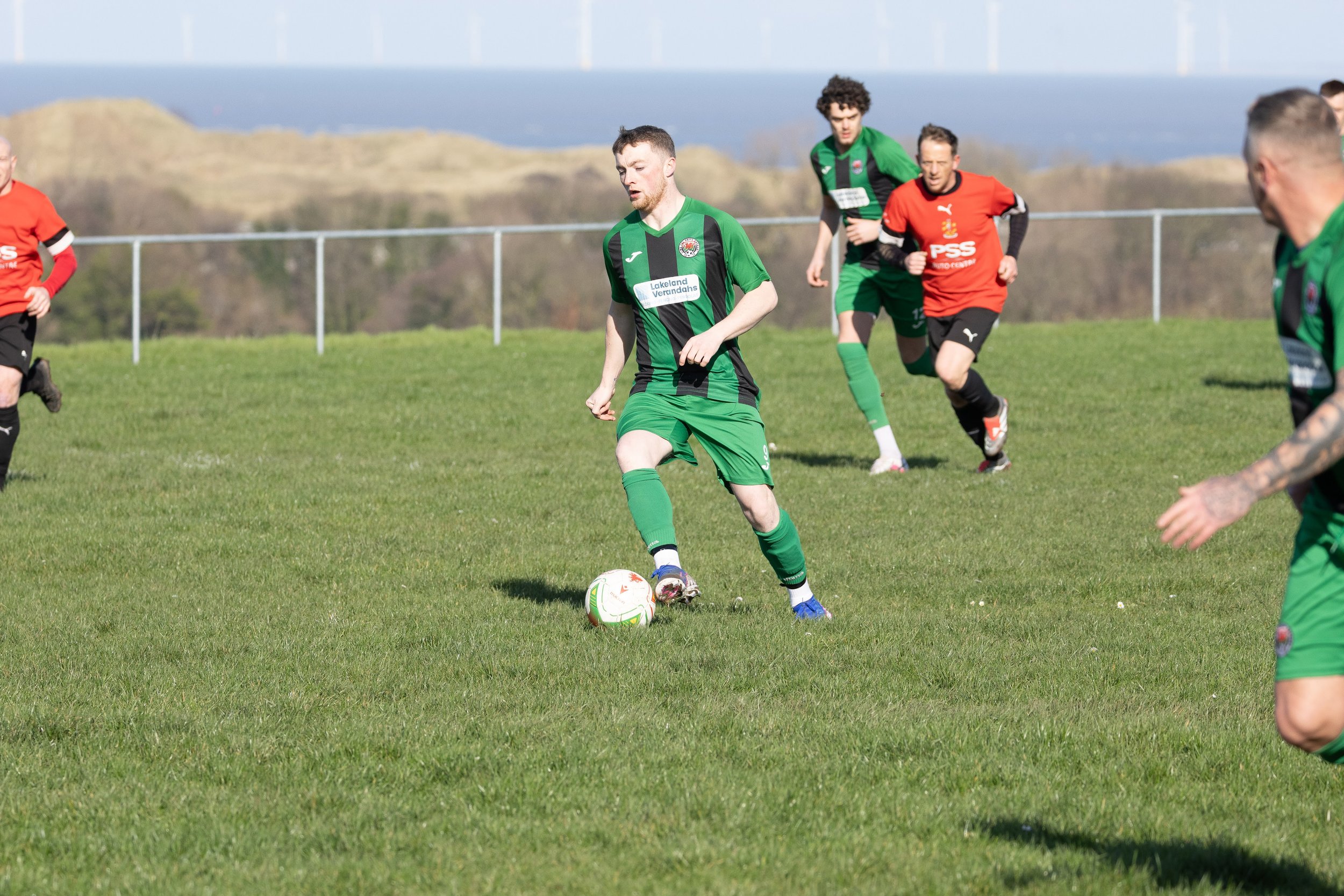 Soccer players on a grassy field, with one player in green controlling the ball while others chase nearby, outdoors with an open landscape in the background.