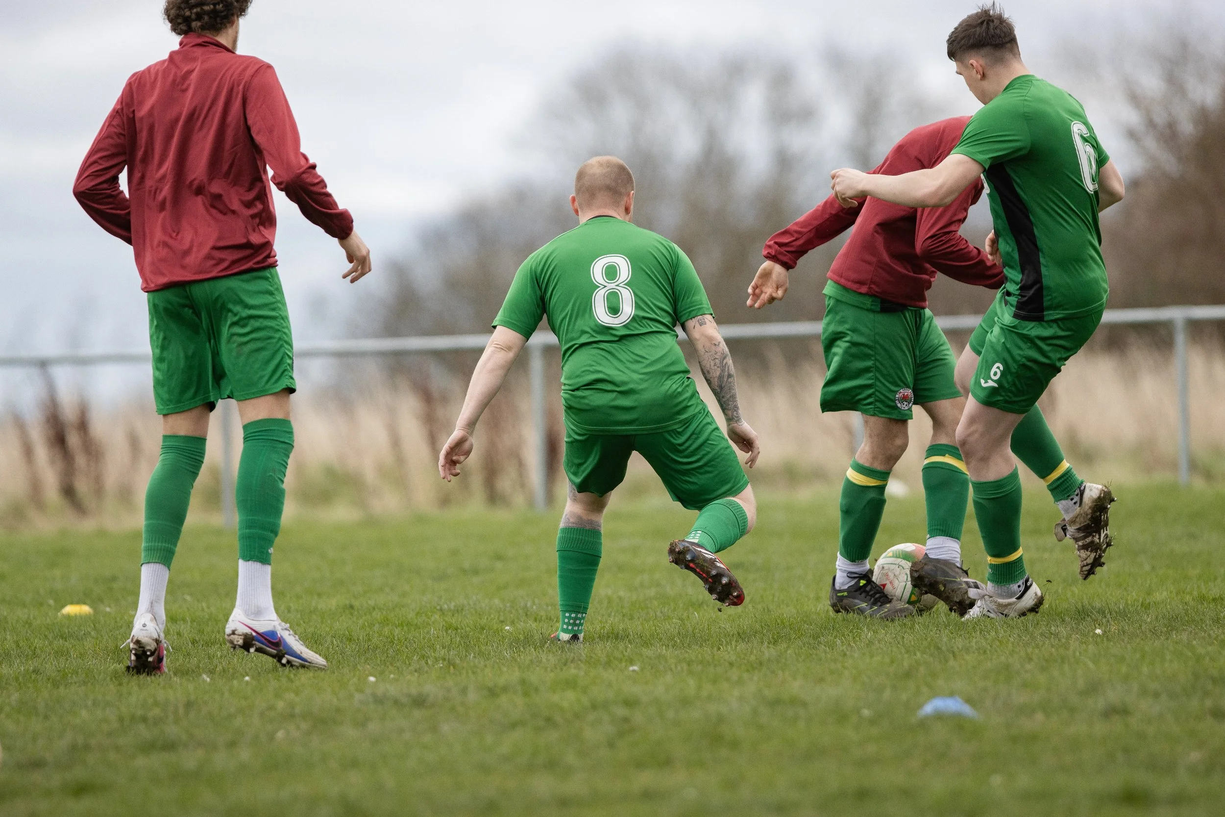 Four soccer players on a field during a game, with one wearing a green jersey with the number 8, actively engaged near the ball, and others in red jerseys.”}
