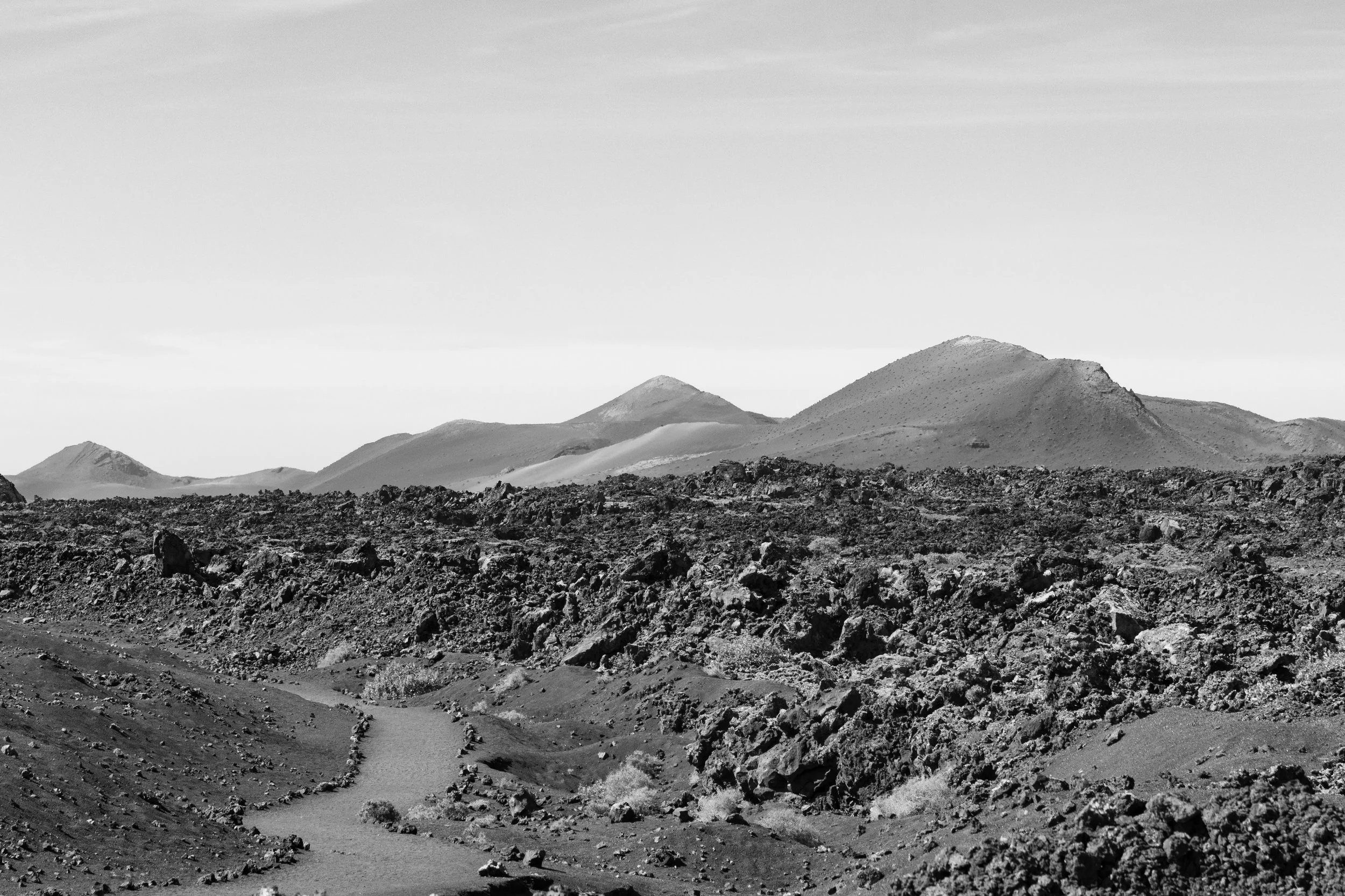 A black and white landscape of volcanic terrain, featuring a rocky foreground, a winding dirt path, and distant volcanic mountains under a clear sky.