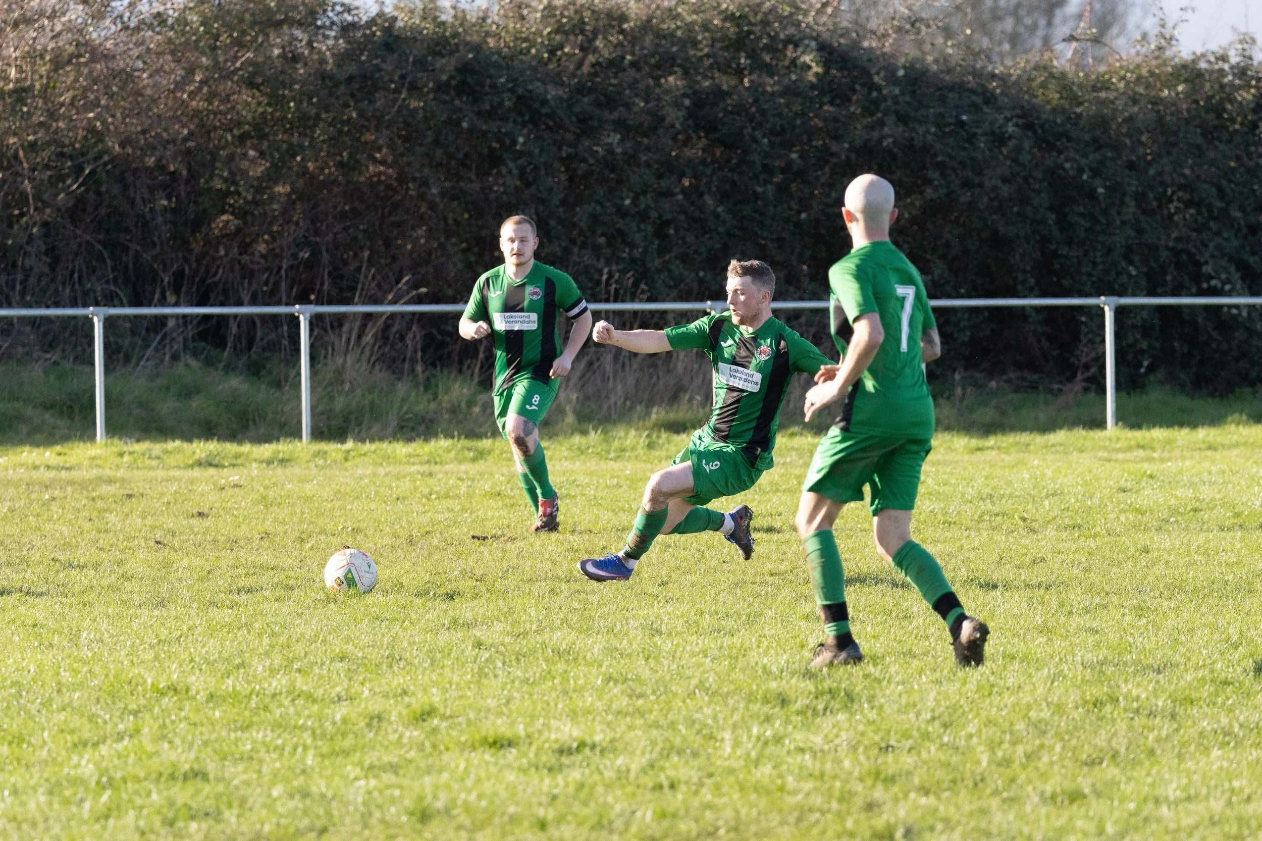 Three soccer players in green jerseys are on a grassy field during a game. One player is kicking the ball, while the other two are nearby, one of whom has a bald head.