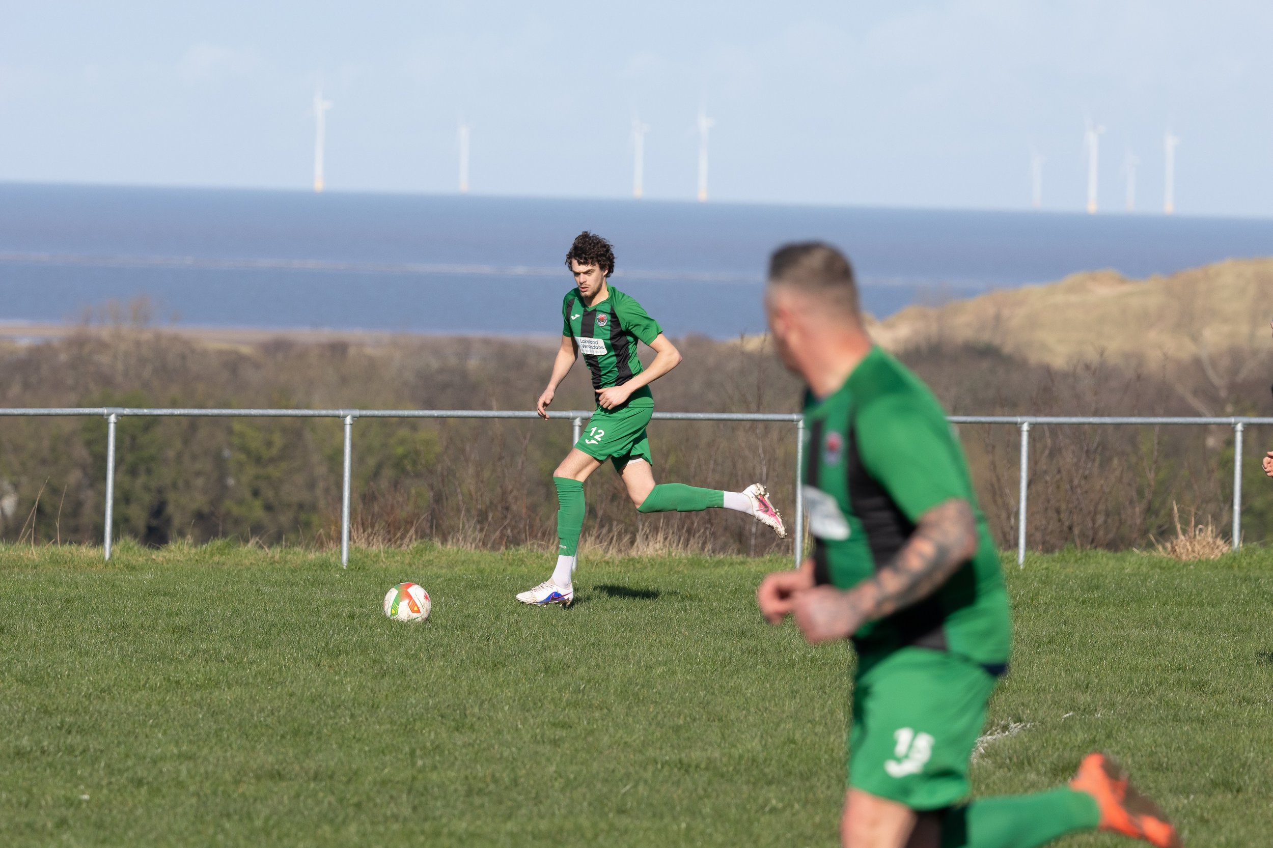 Soccer players in green uniforms practicing on a grassy field with a landscape of hills and wind turbines in the background.
