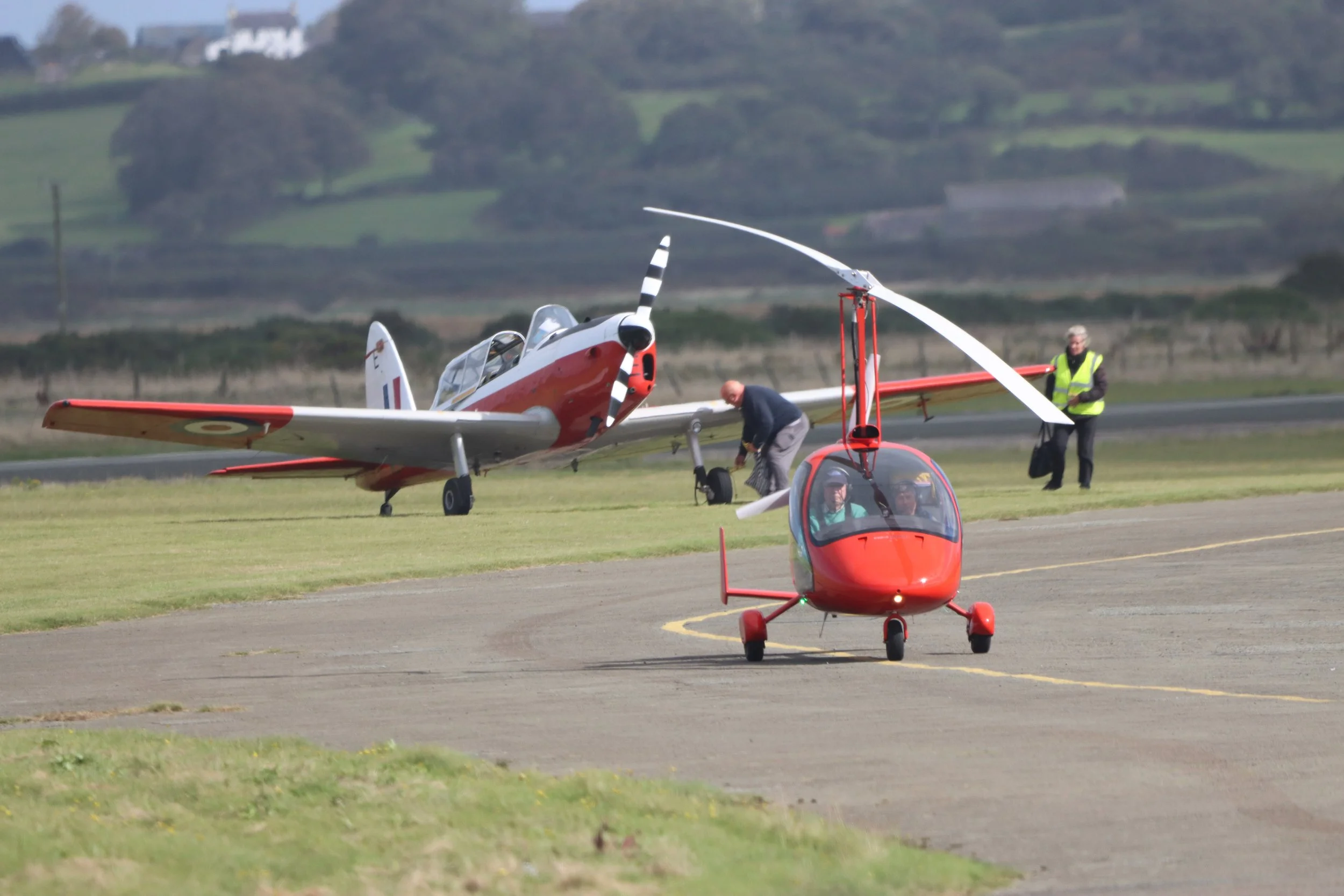 A small red helicopter and a vintage propeller airplane are on a grassy airstrip, with people preparing the aircrafts for takeoff and a man in a high-visibility vest standing nearby. The background features rolling green hills.