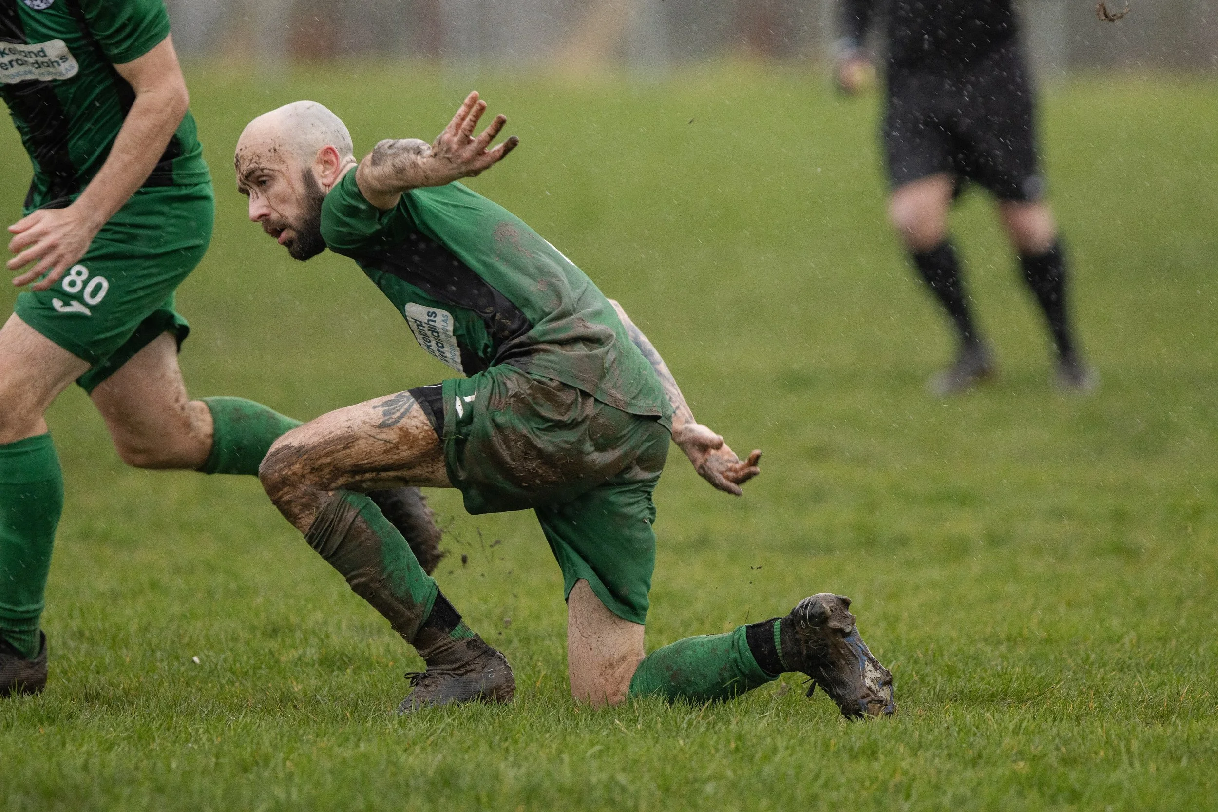 A soccer player in green uniform falling and grabbing his ankle during a rain-soaked match.