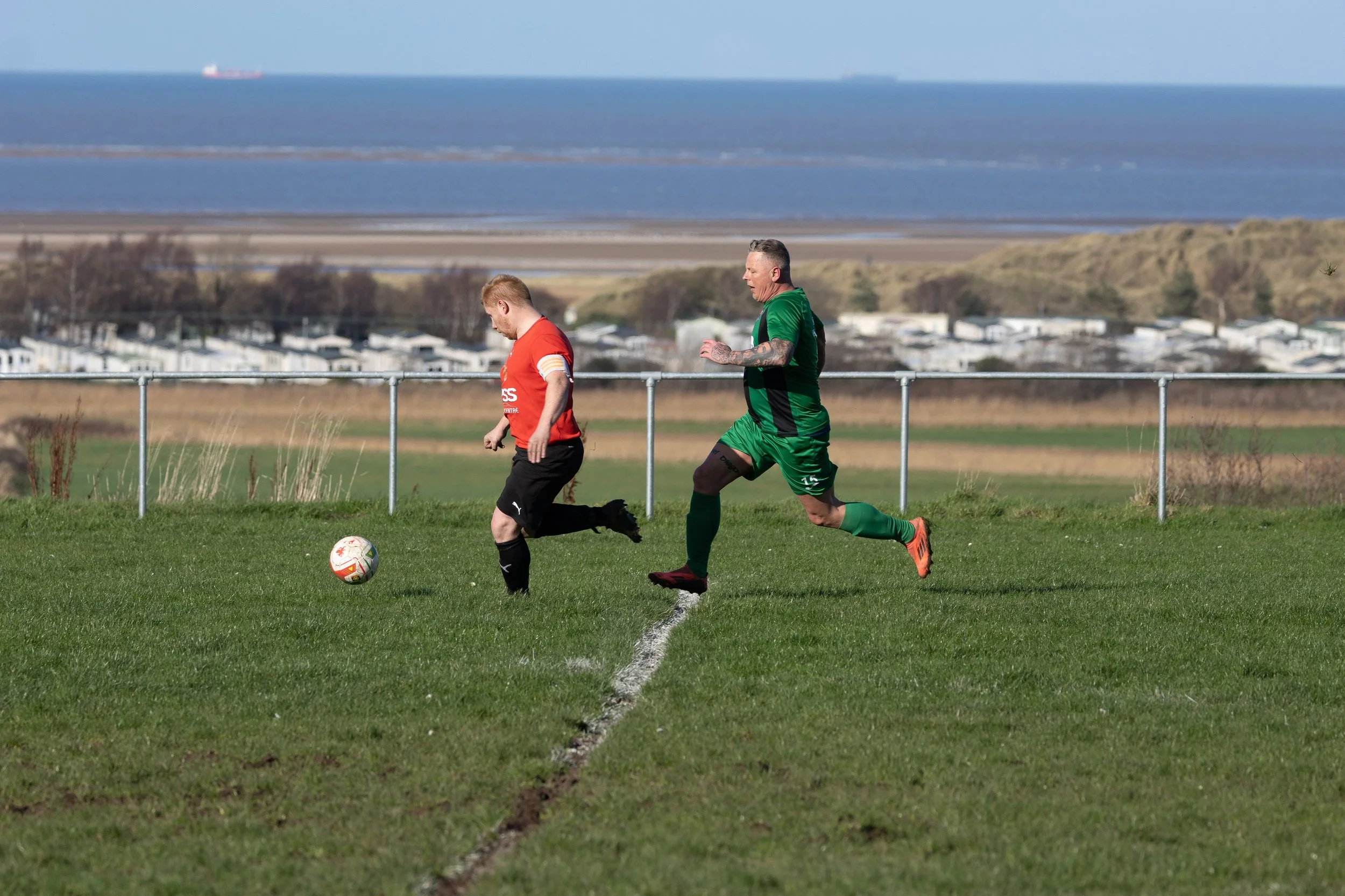 Two soccer players, one in red and black and the other in green, chase a soccer ball on a grassy field with a coastal landscape and housing in the background.