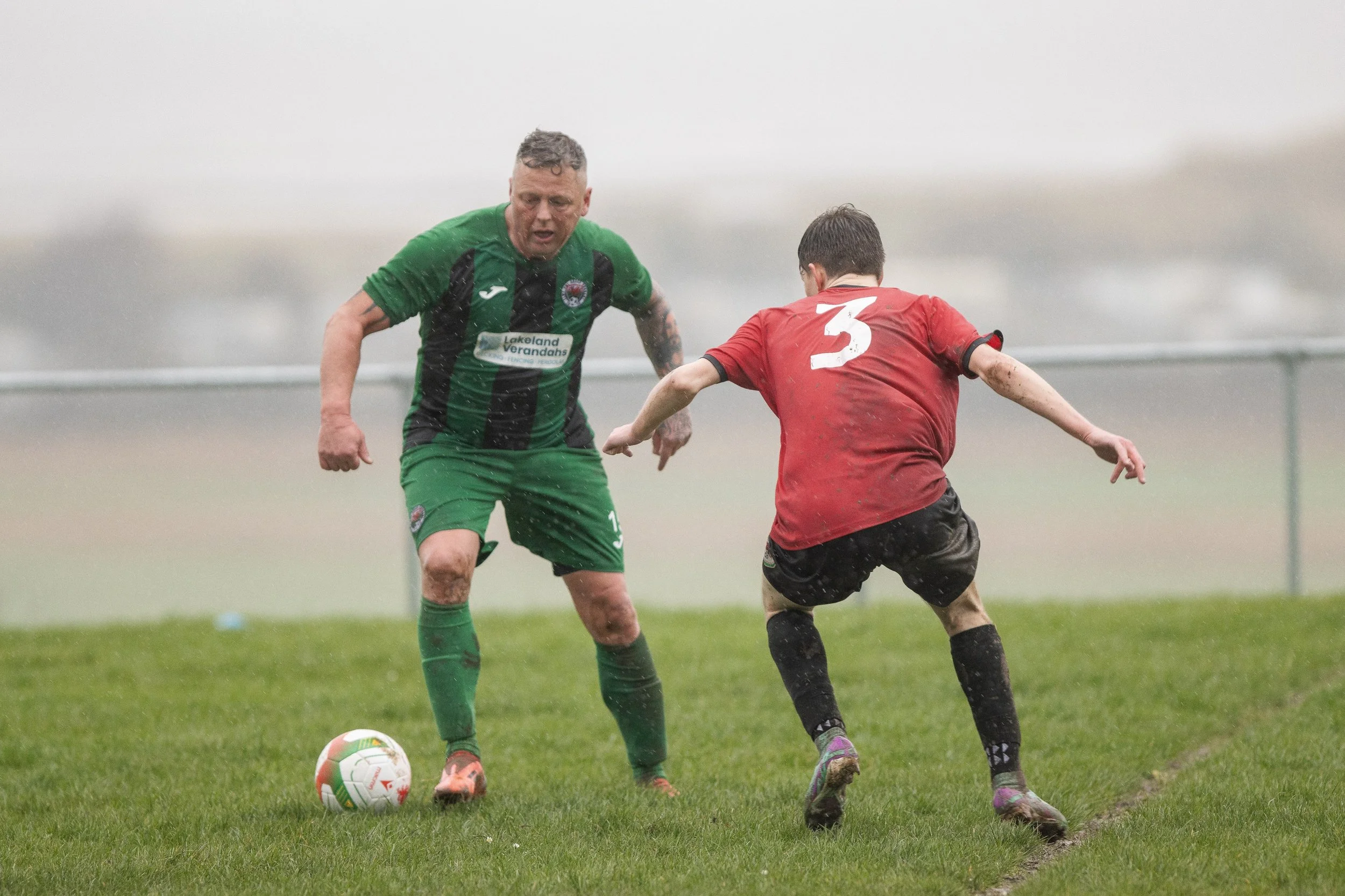 Two soccer players compete for the ball on a rainy field, one in a green and black uniform, the other in a red uniform with the number 3.