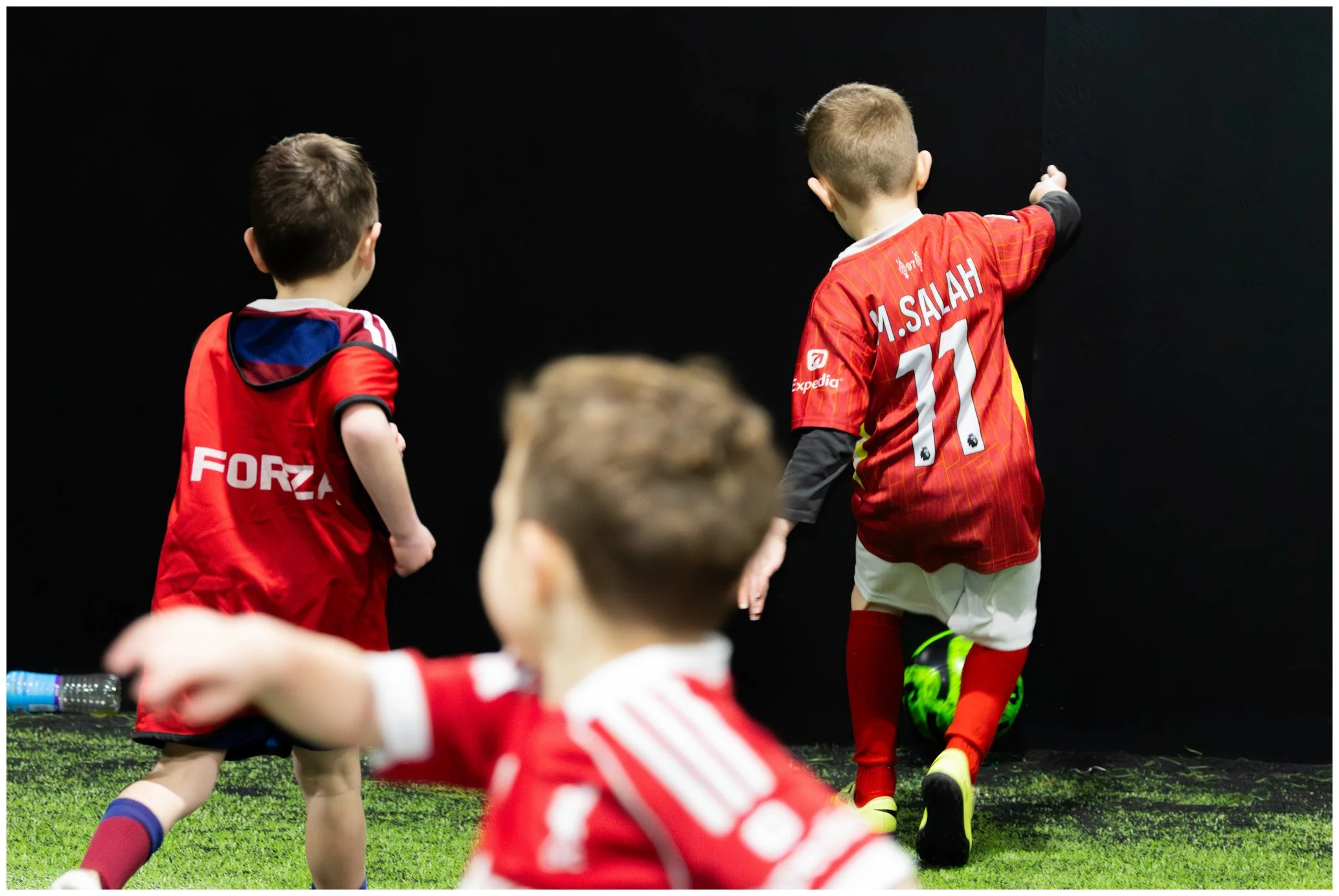 Three young boys in red soccer uniforms on an indoor field, with a black wall in the background. One boy is facing away, pointing towards the wall, while another boy in the foreground is out of focus. The third boy is partially visible, reaching out 