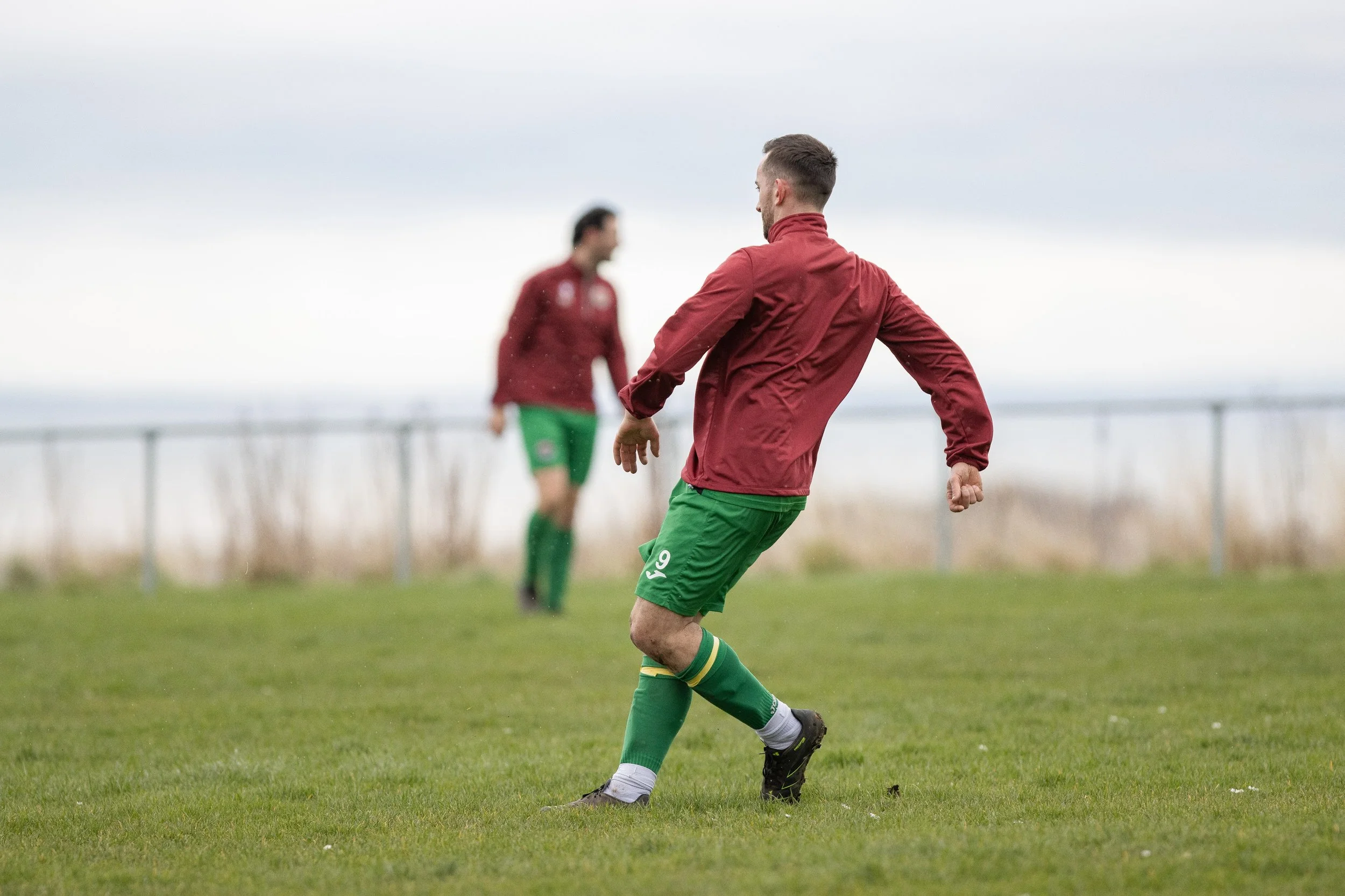A soccer player in a red jacket and green shorts kicks a ball on a grassy field, with another player in the background