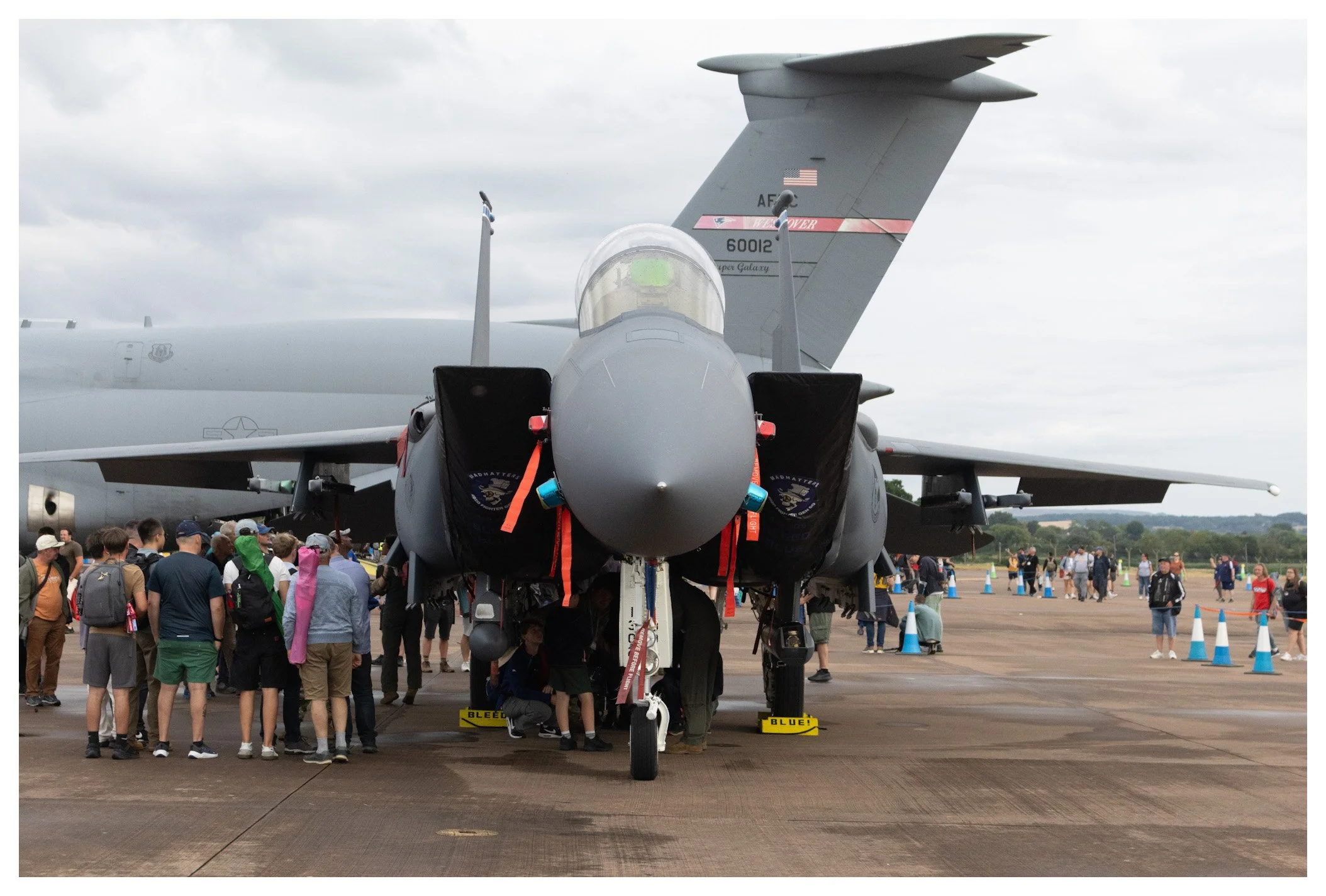 Front view of a military fighter jet on display at an airshow, with a crowd of people observing around it and blue traffic cones in the background.